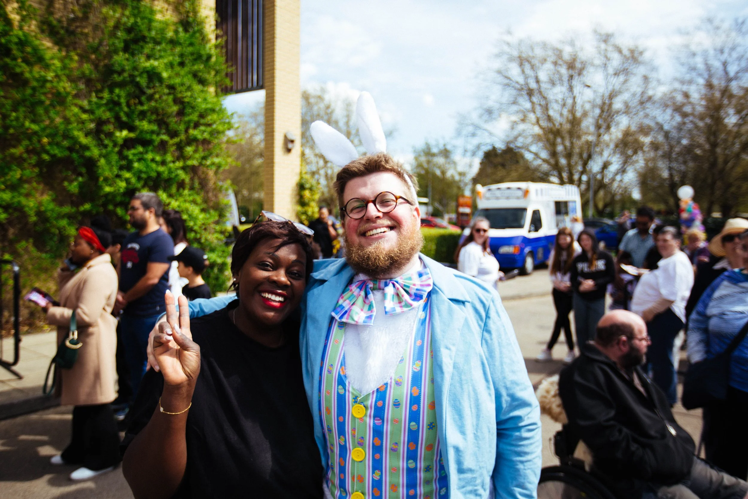 People celebrating outdoors, with one person dressed as an Easter Bunny, he has white bunny ears, a colorful bow tie, and a pastel-colored vest. Someone is making a peace sign, and everyone is smiling. The background includes trees, a building, a bus