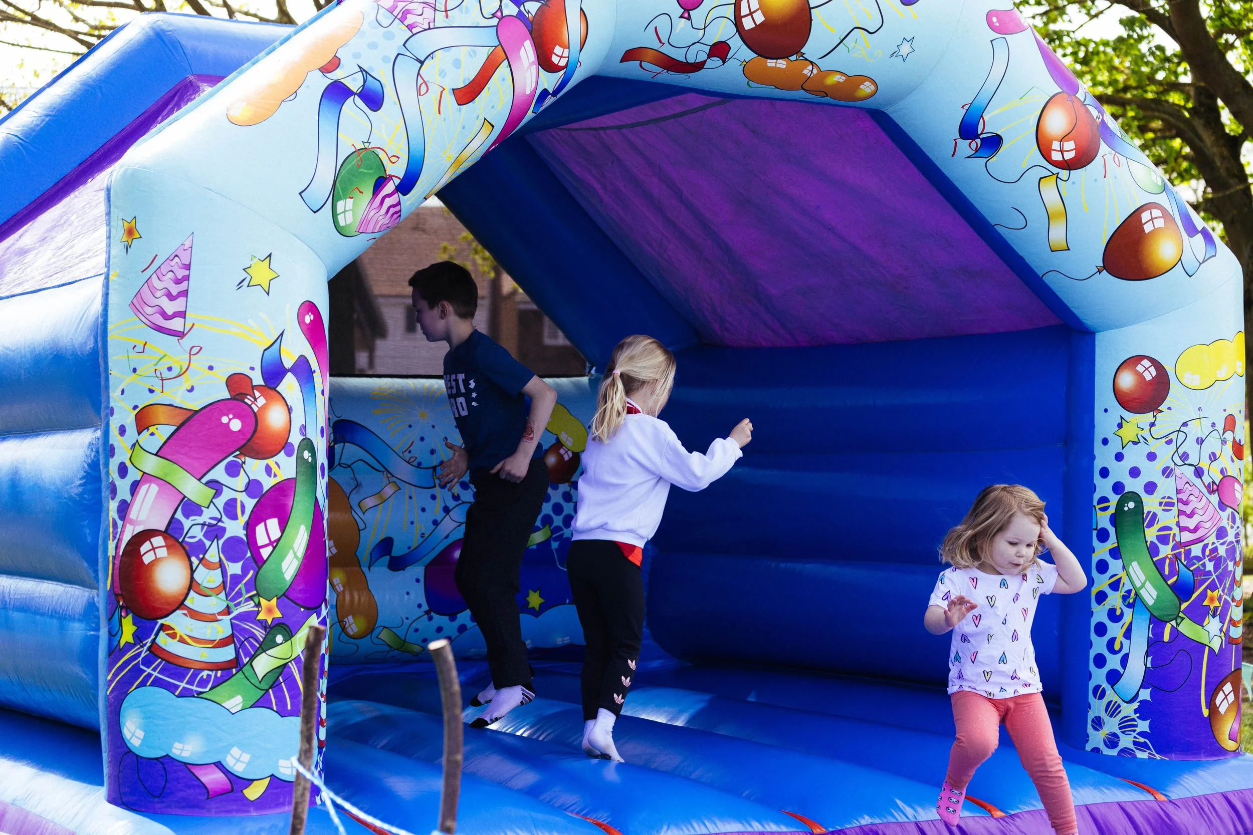 Children playing on a colorful inflatable bounce house decorated with balloons, party hats, and streamers during daytime outdoors.