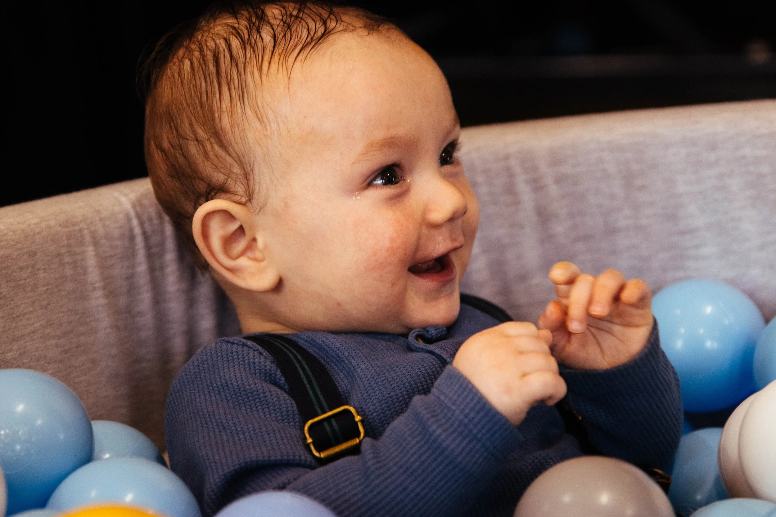 A young child with wet hair smiling and playing in a ball pit with blue, yellow, and white plastic balls.