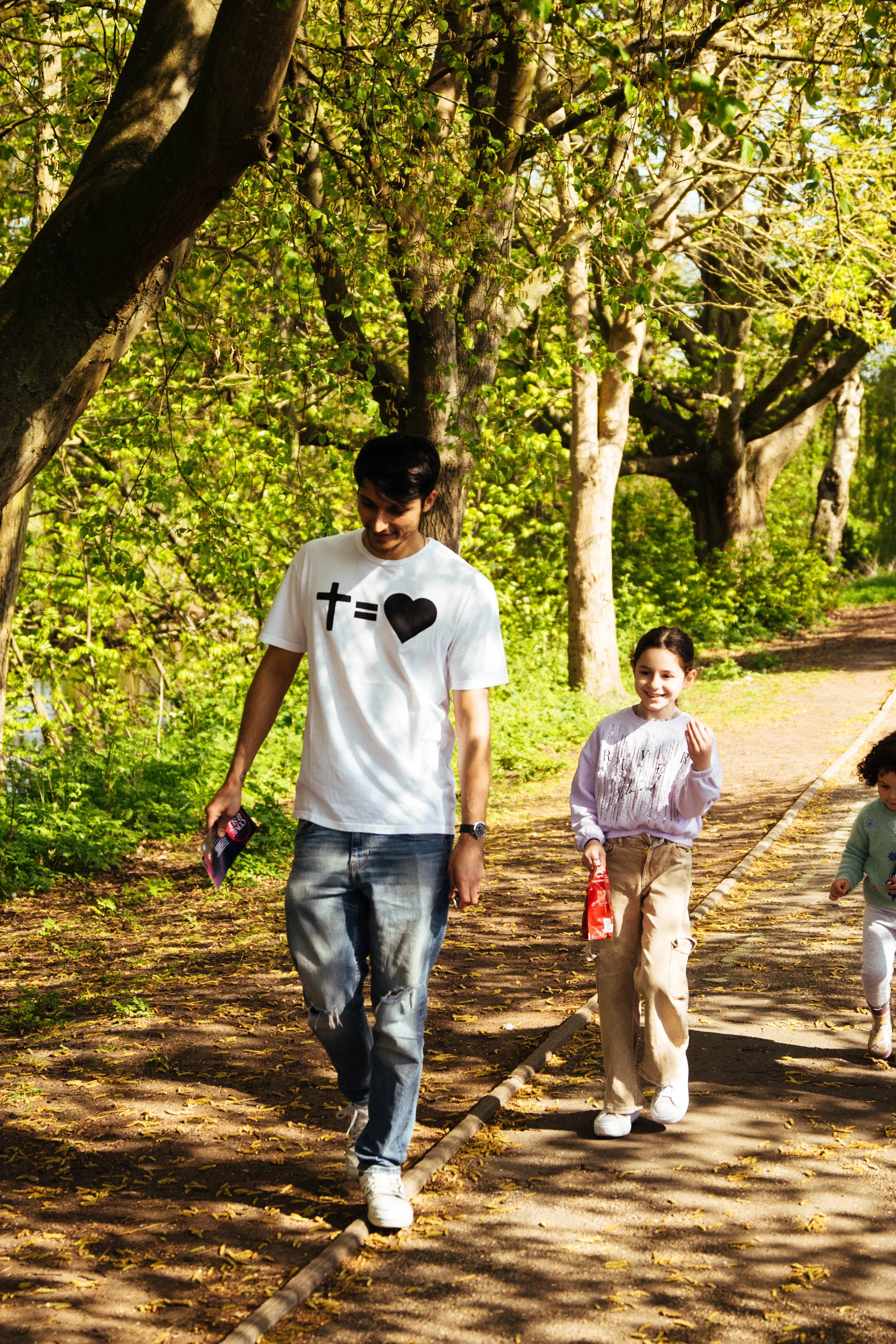 A man and two young girls walking along a tree-lined path outdoors on a sunny day.