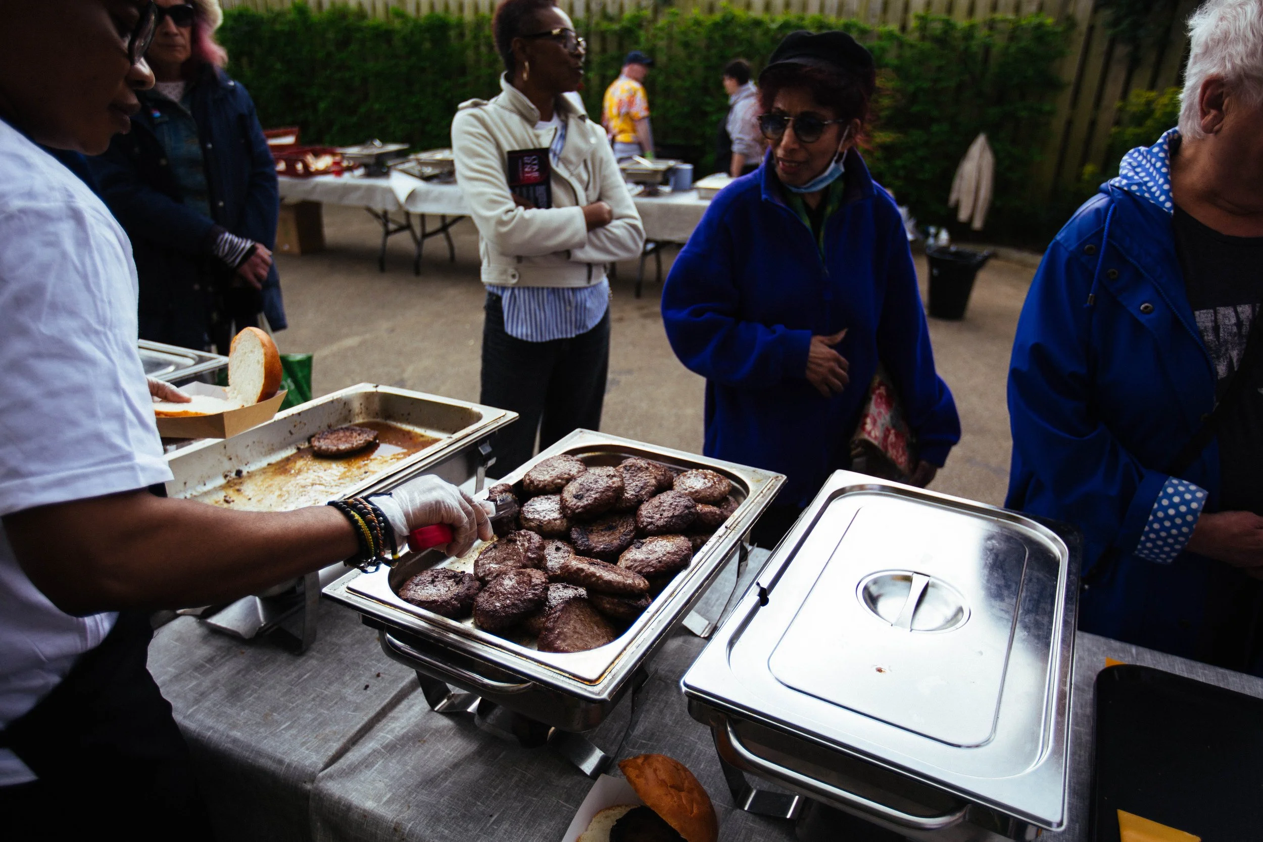 People standing in line at outdoor food buffet, where a person is serving cooked meat patties.