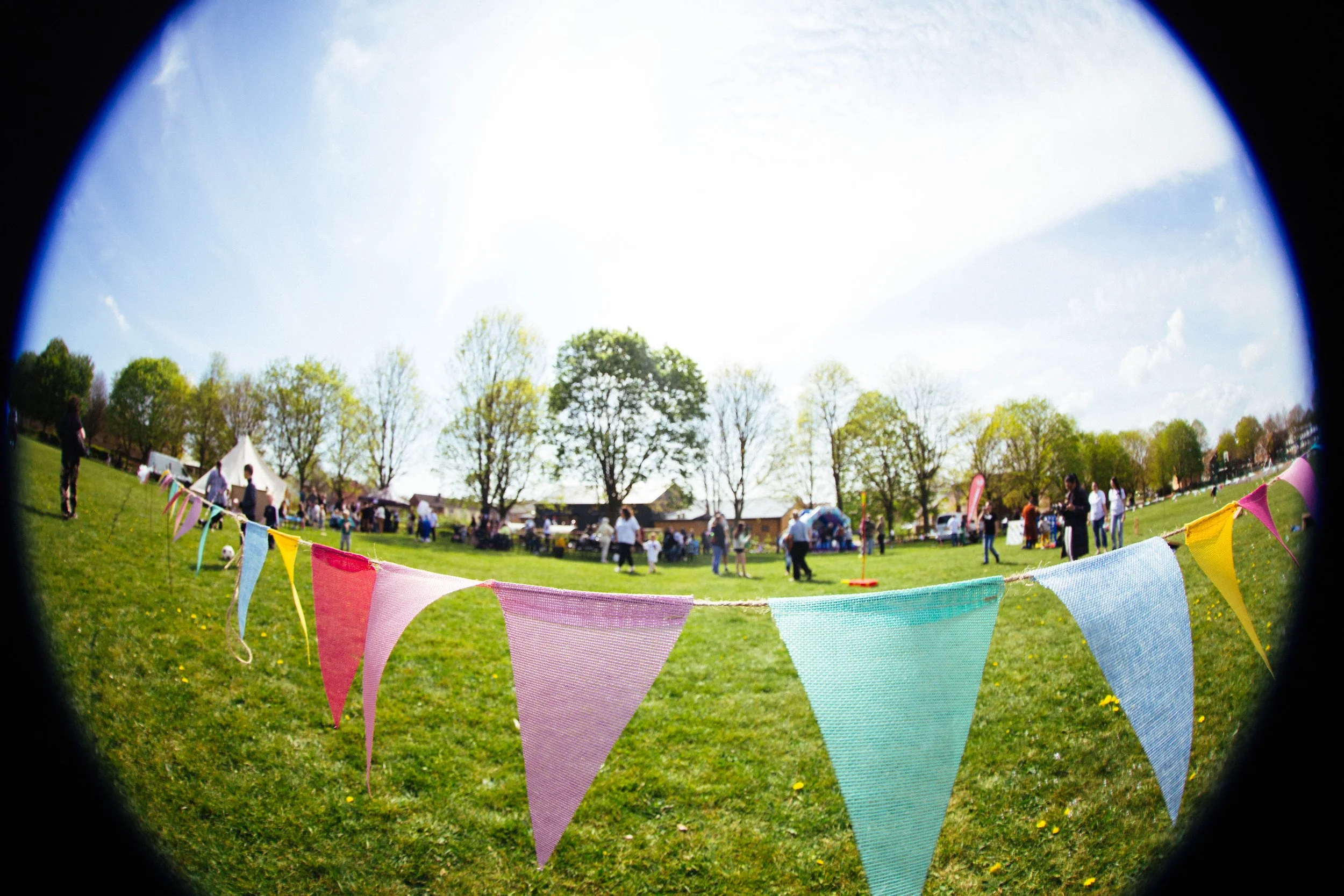 Outdoor park scene seen through a fisheye lens with colorful bunting in the foreground, people gathered, tents, trees with green leaves, and a blue sky with some clouds.
