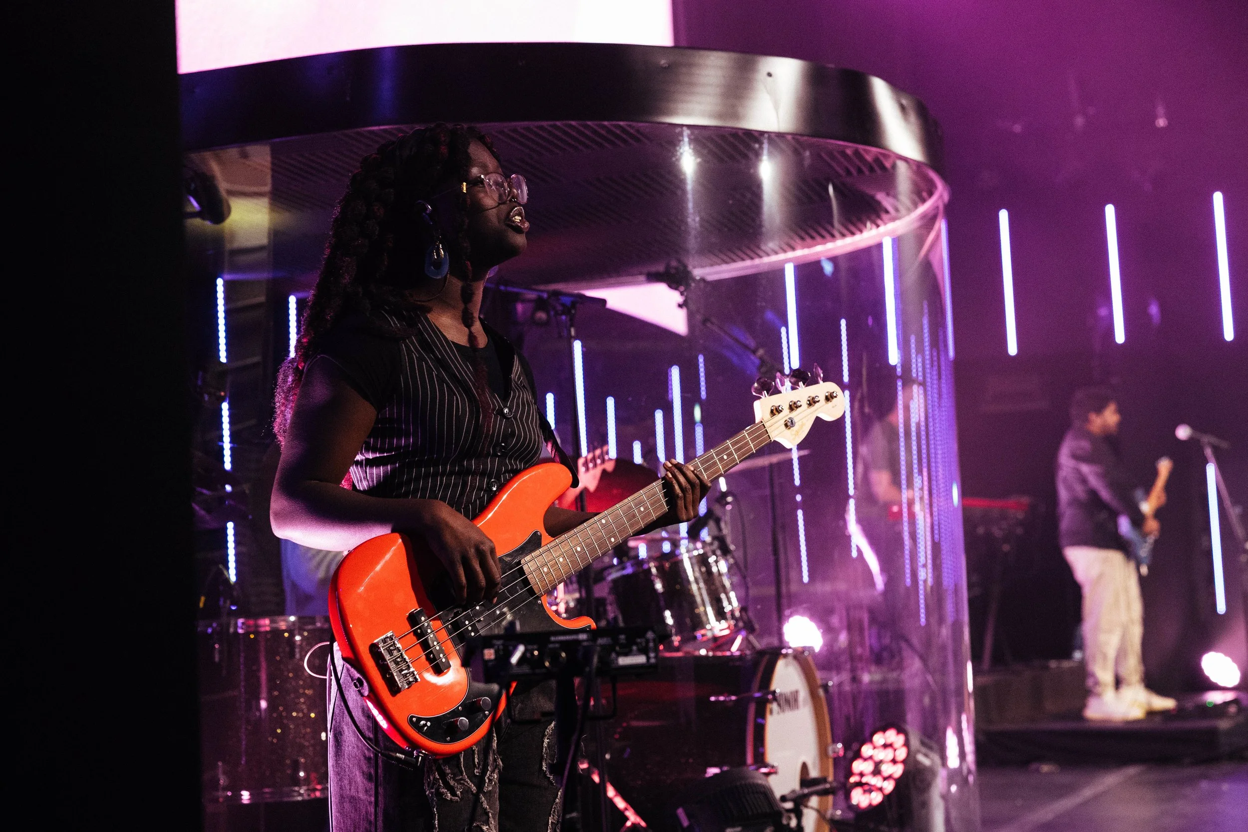 A female musician playing an orange electric bass guitar on stage during a concert, with purple and pink lighting, and other band members in the background.