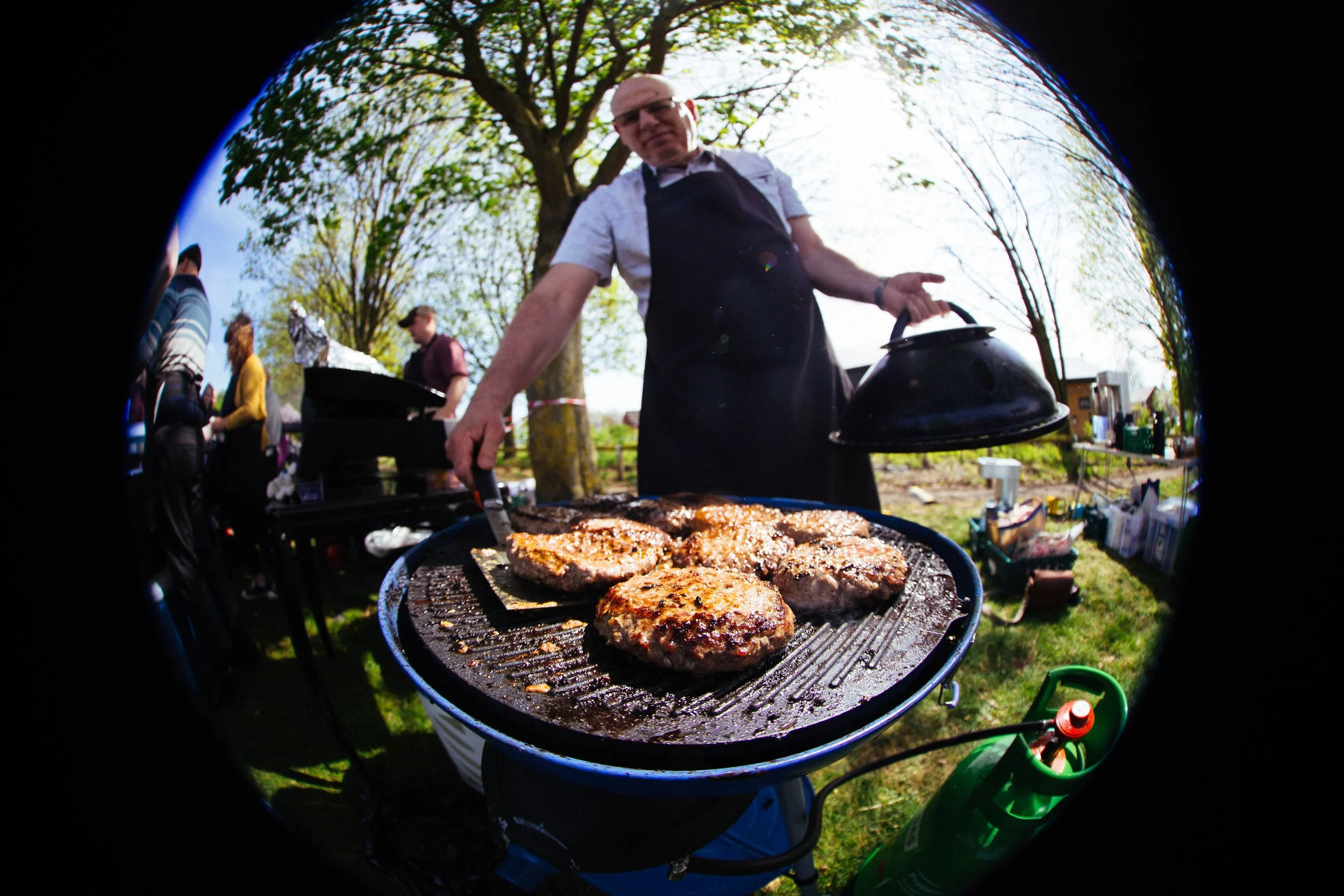 Man grilling hamburgers outdoors during sunny day, people in the background, trees and grass visible.