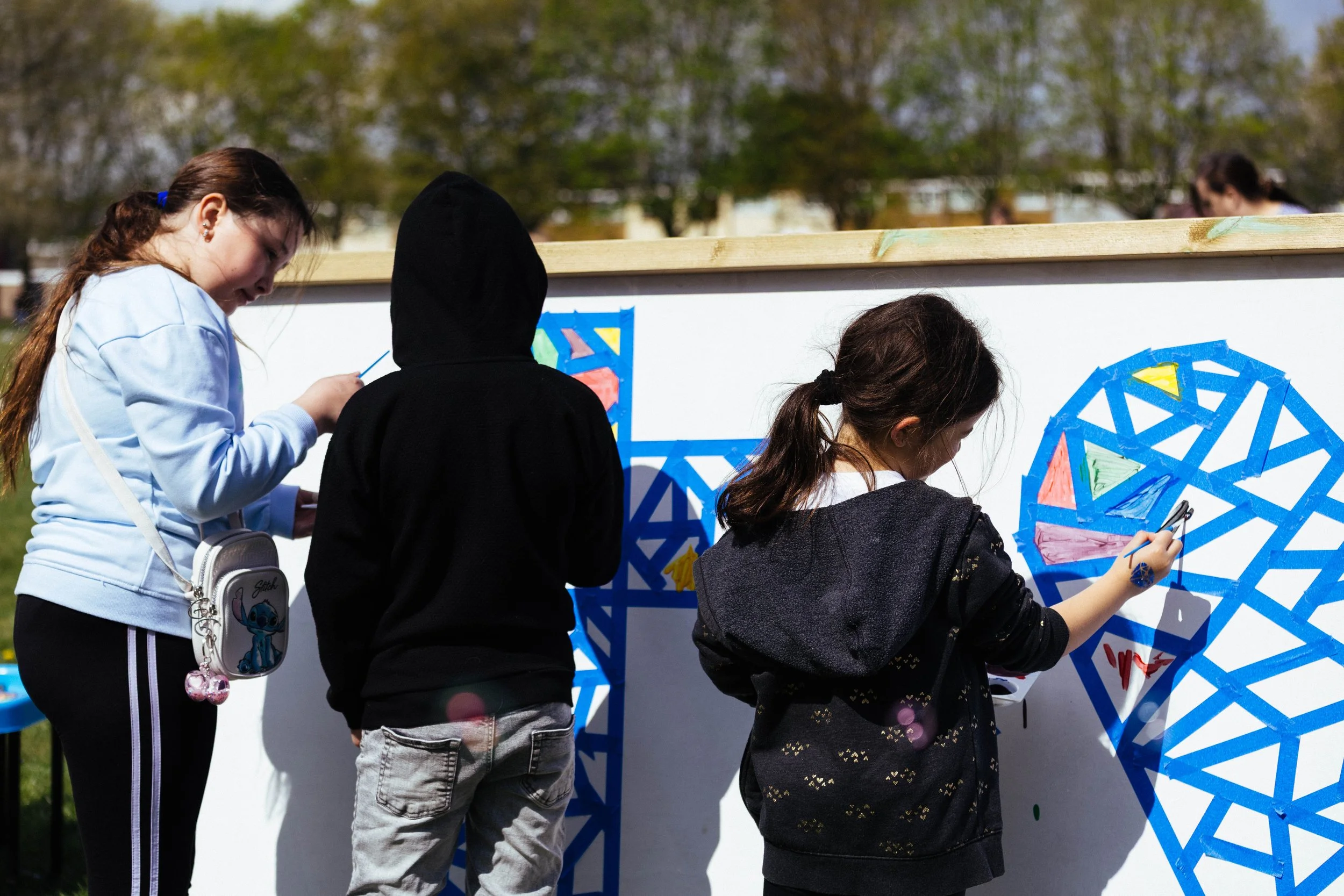 Three children are painting a large white board outdoors, with trees and other people in the background.