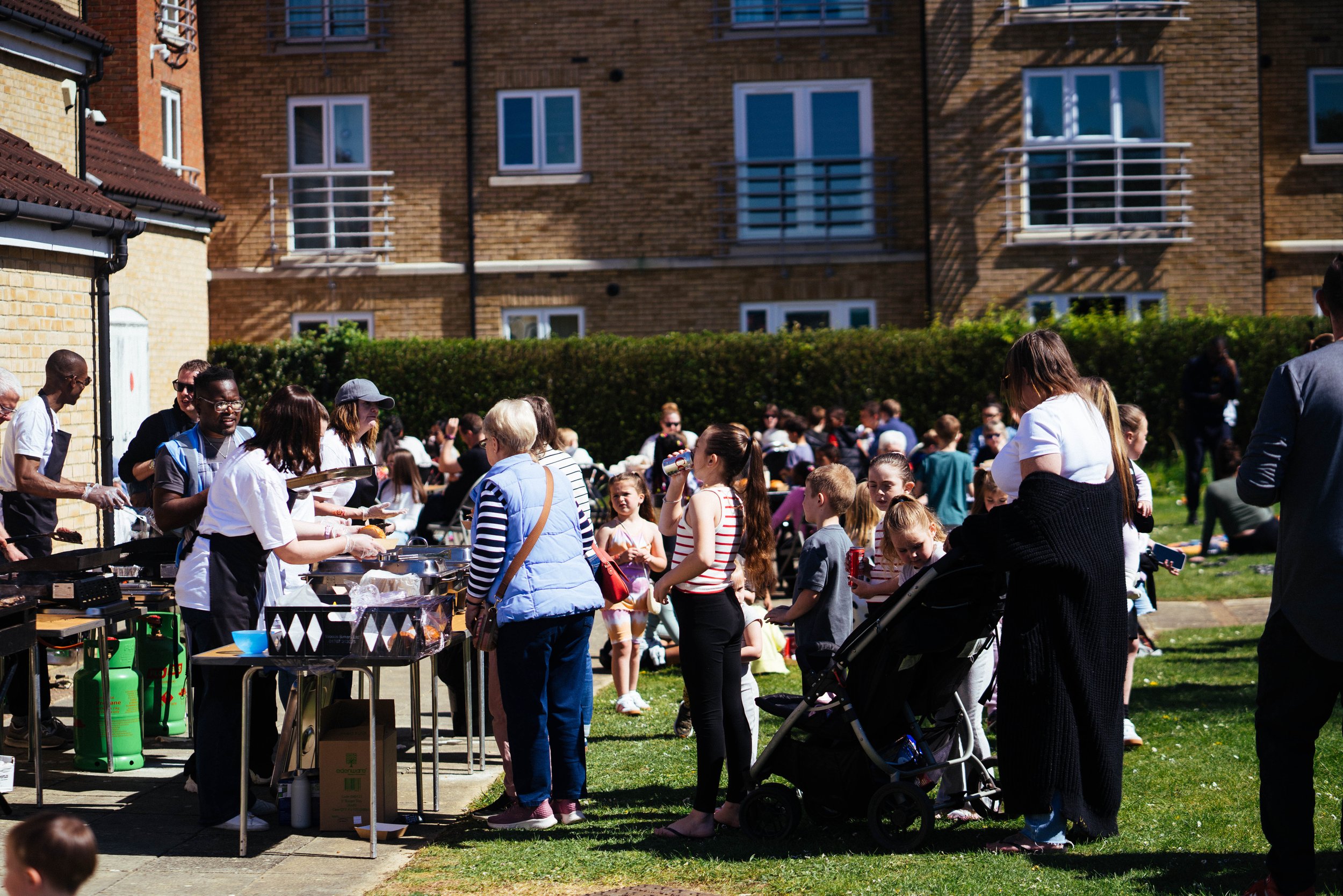 People at an outdoor community gathering or barbecue, with children and adults standing in line for food, servers preparing food, and a grassy area with more people in the background.