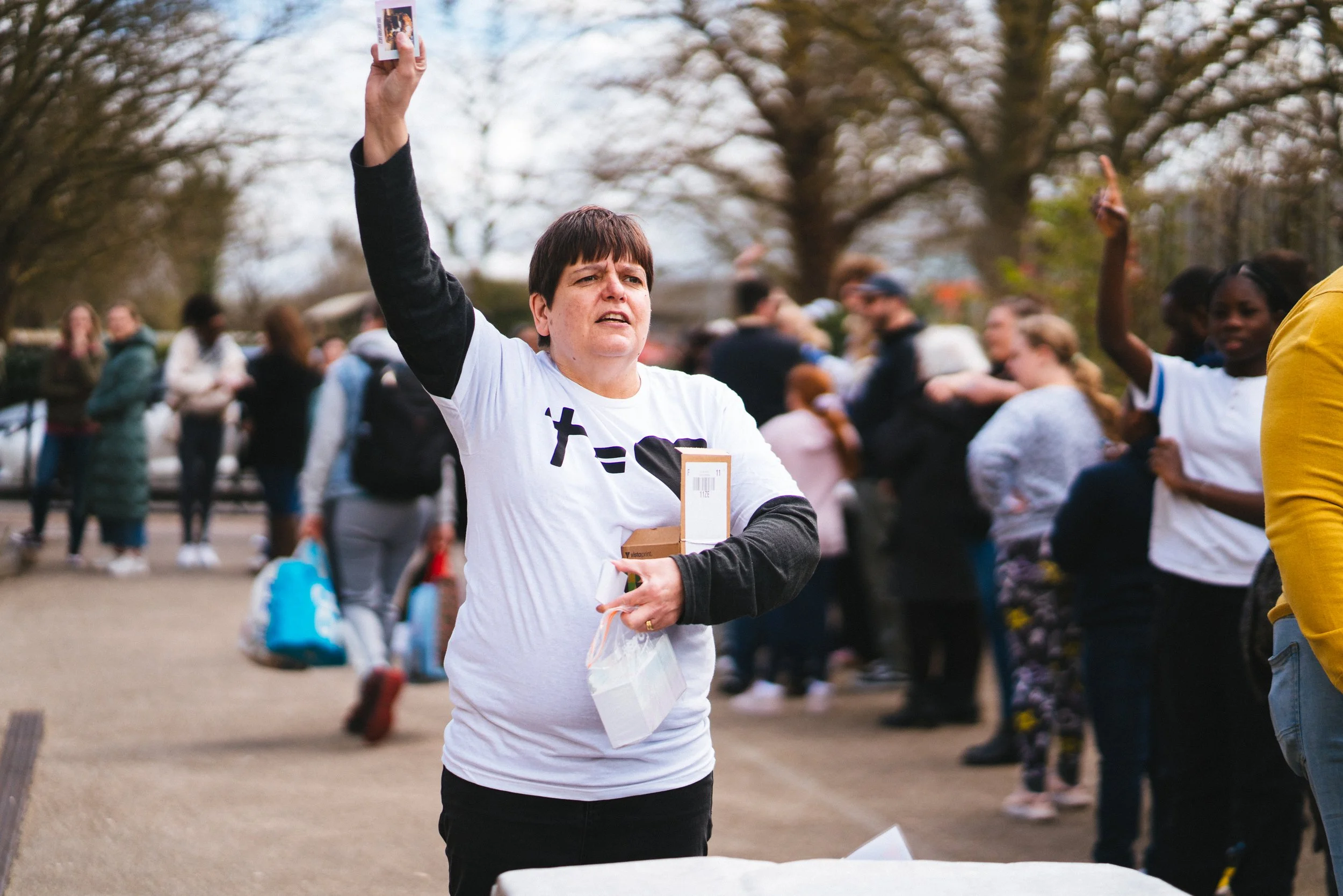 A woman with short dark hair wearing a white T-shirt with black sleeves and black pants stands outdoors at a gathering, holding a phone in the air and a box with a paper bag in her other hand. Other people are in the background near trees, some raisi