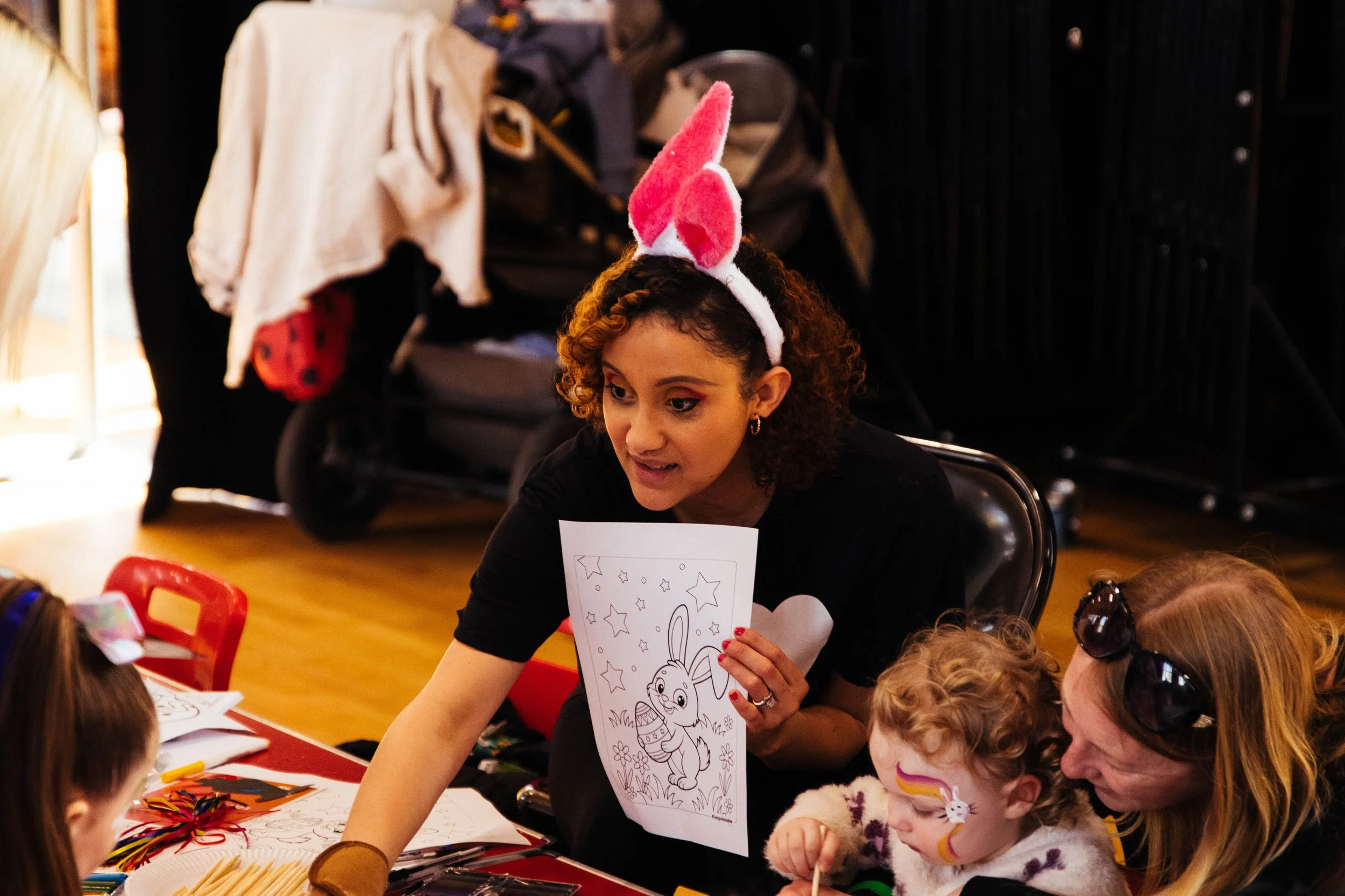 Woman with curly hair wearing pink bunny ears headband holding a coloring sheet with a bunny, surrounded by children at a craft table, some with painted faces, in a festive indoor setting.