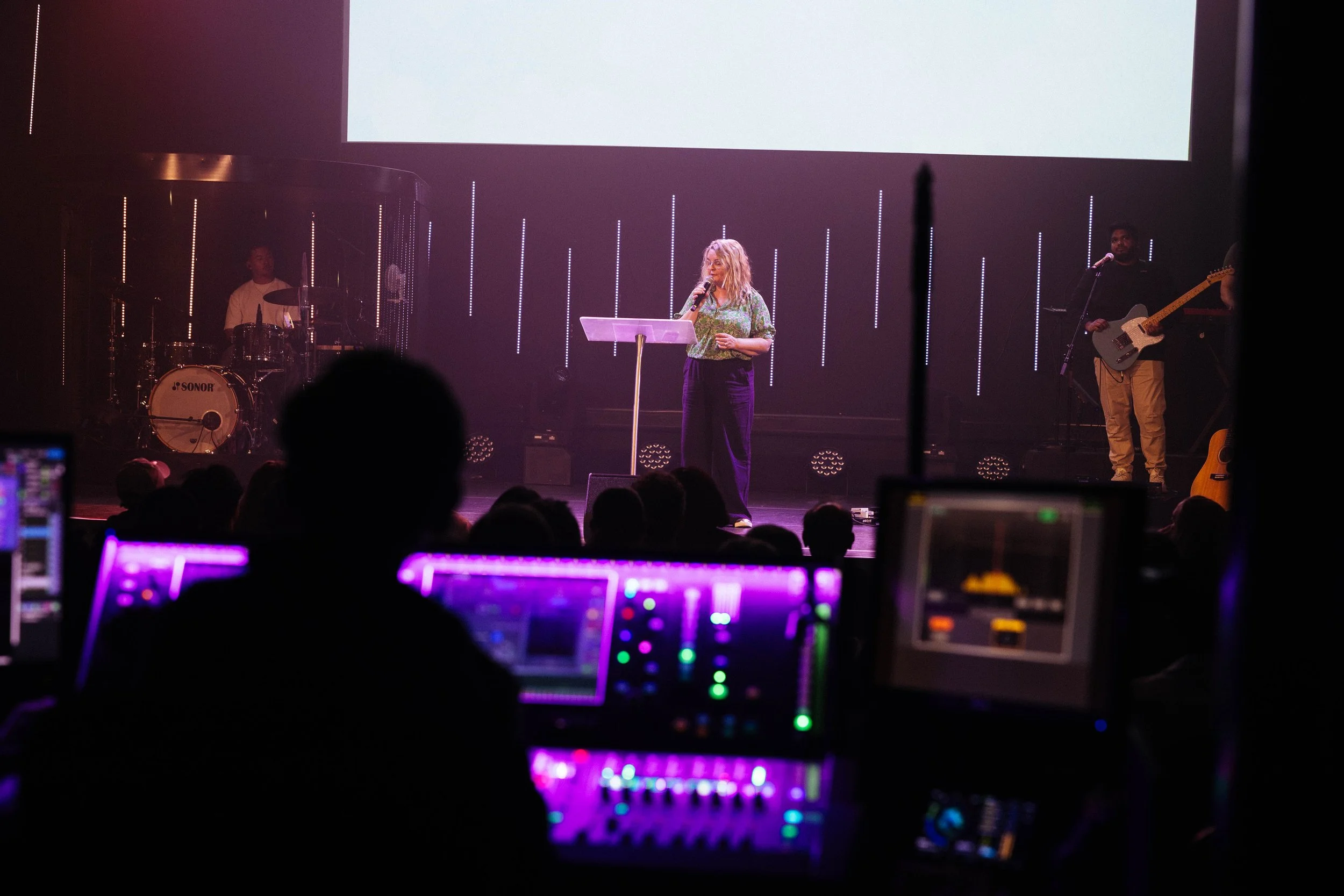 A woman speaking on stage with a microphone, accompanied by a band with a drummer and a guitarist, during a performance or presentation. The audience and sound equipment are visible in the foreground.