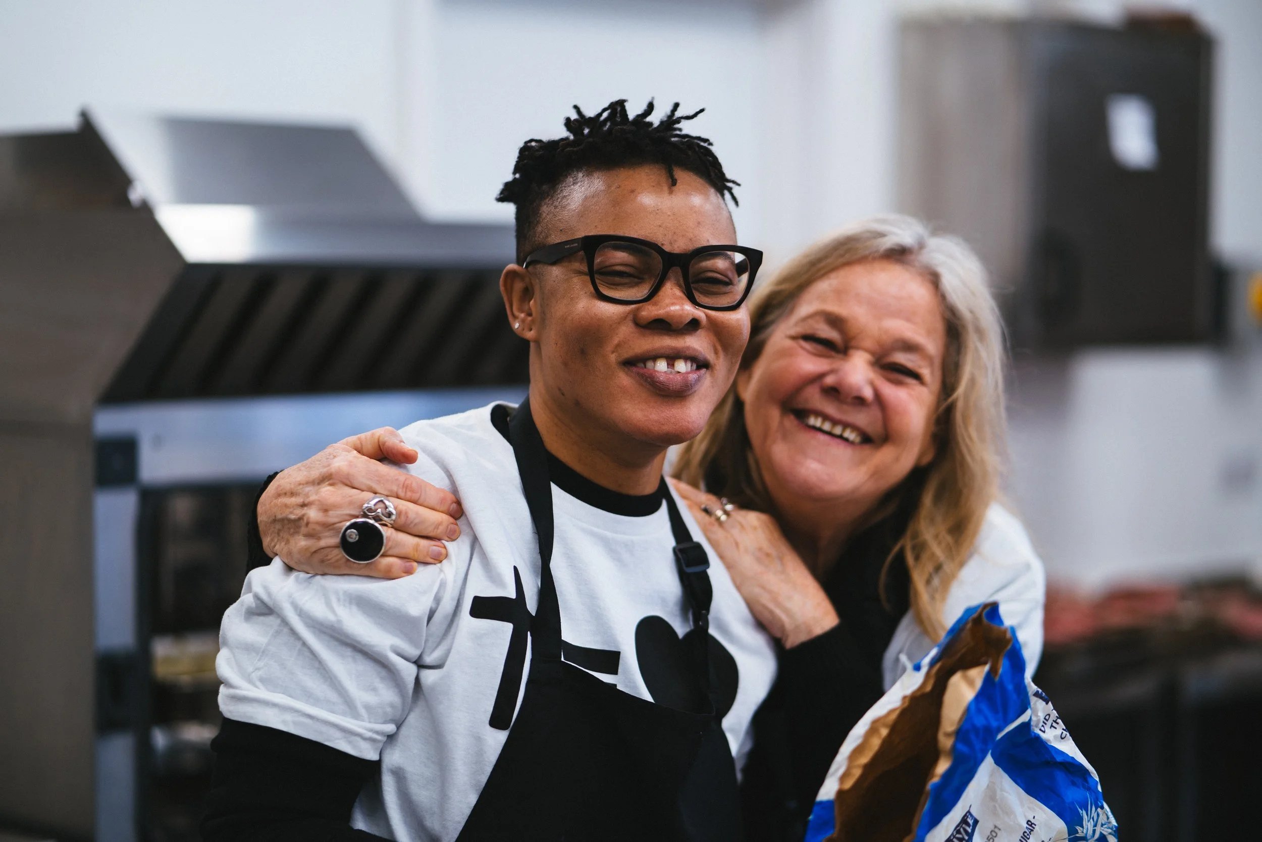 Two women smiling and hugging in a kitchen, one wearing glasses and a black apron, and the other smiling behind her.