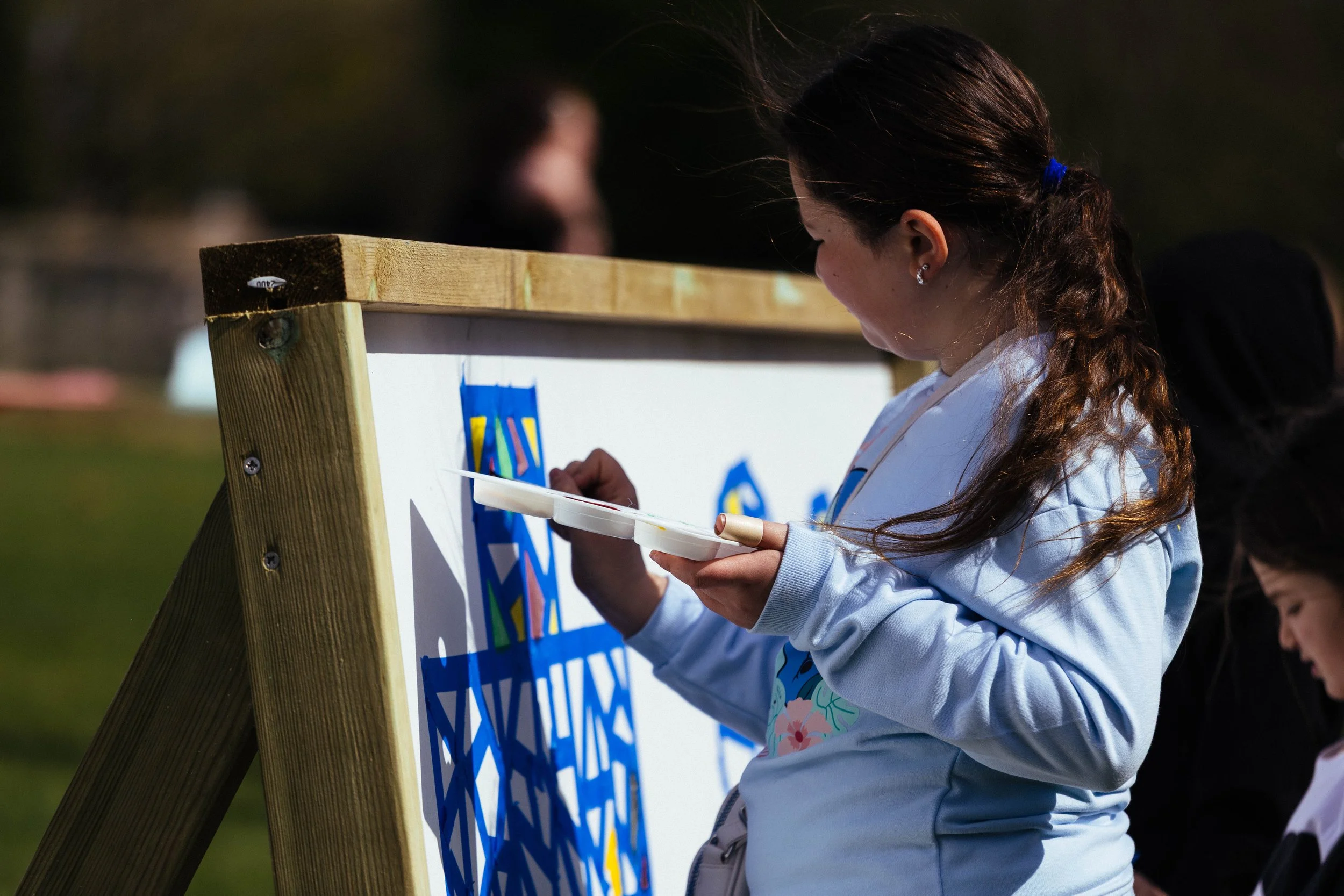 A young girl painting on an outdoor wooden easel with an abstract colorful design, other children are visible in the background.