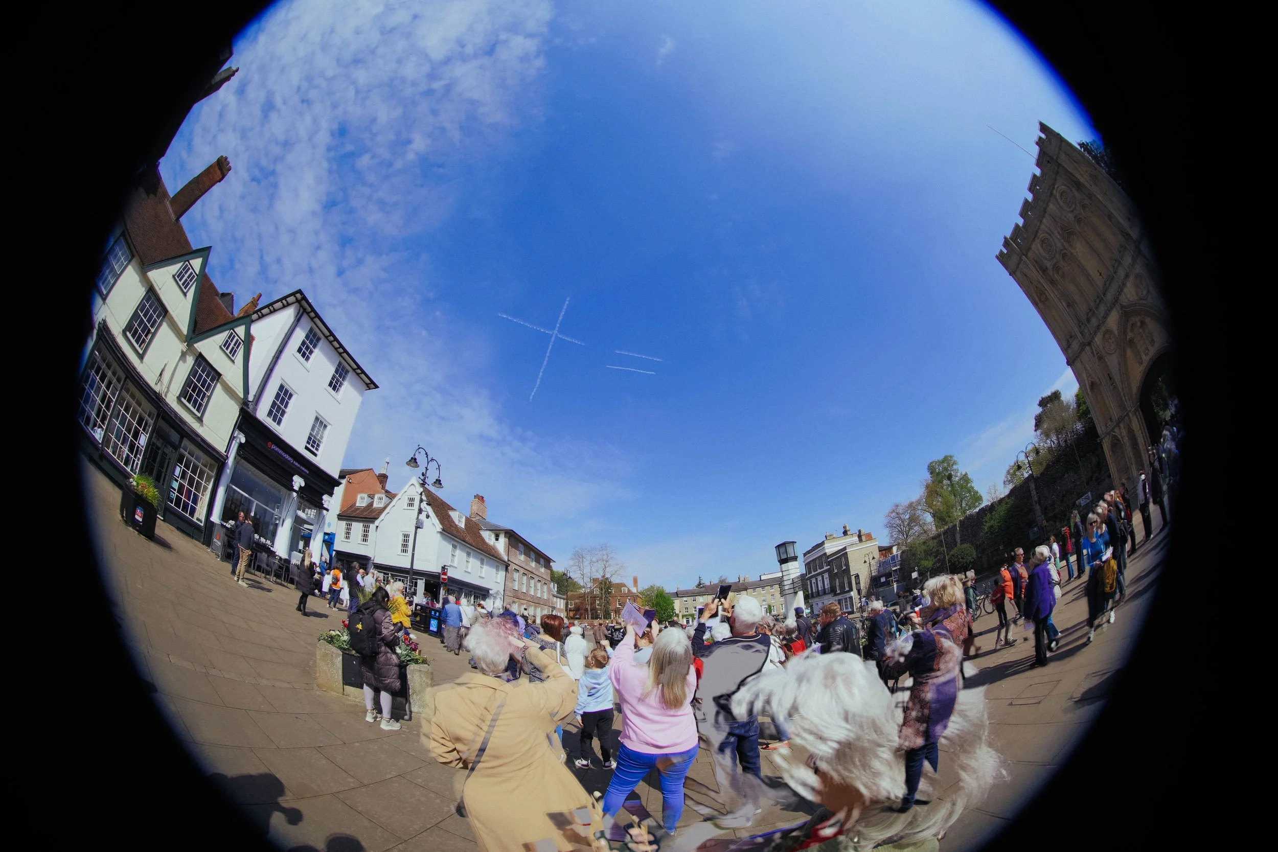 People gathered in a busy town square with historic buildings, a clear blue sky with cloud patterns and contrails, and a large archway on the right side.