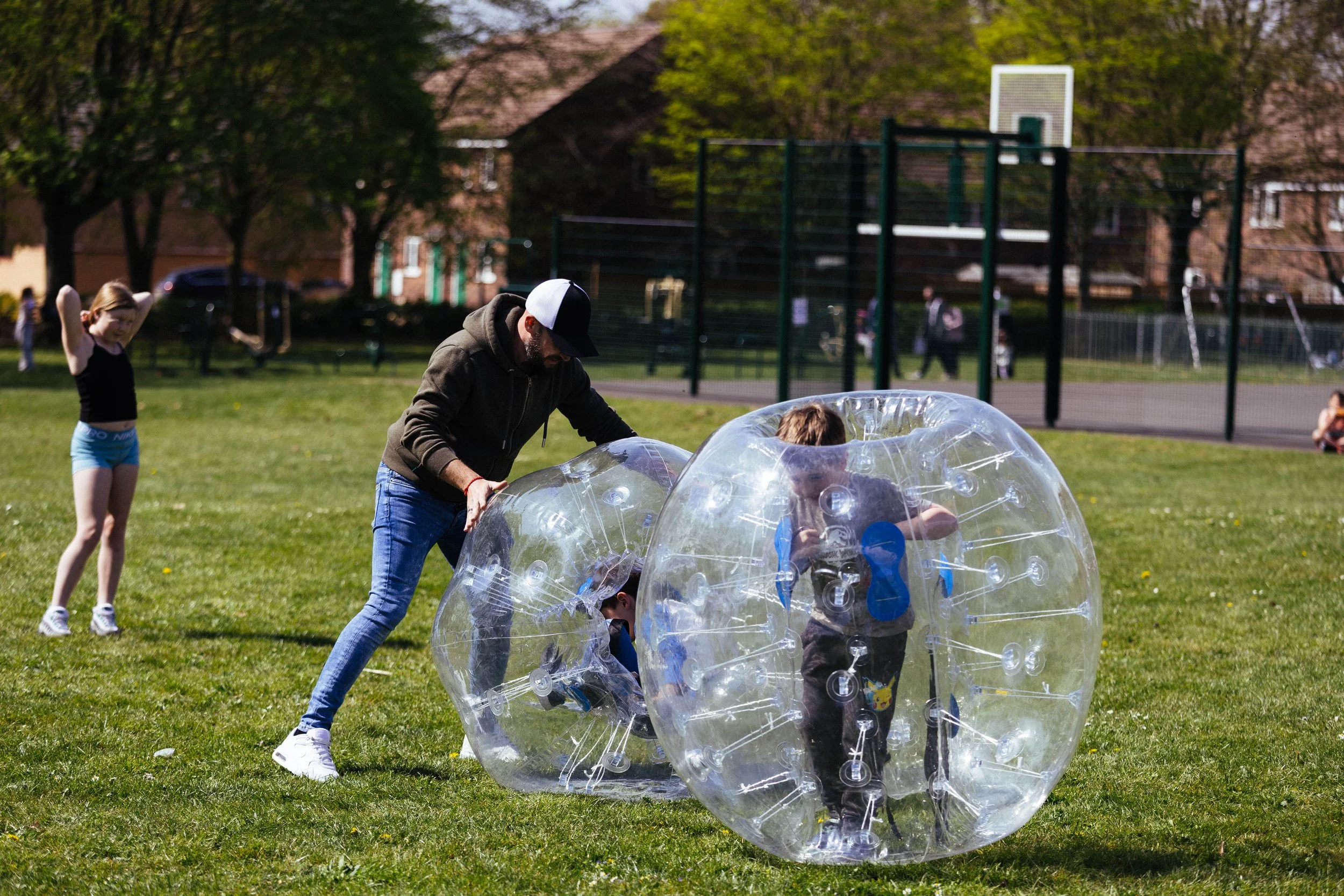A man and two children playing with inflatable zorbing balls on a grass field in a park. The man is helping one child inside a large, transparent inflatable ball, while another child stands nearby. In the background, there are trees, a basketball cou