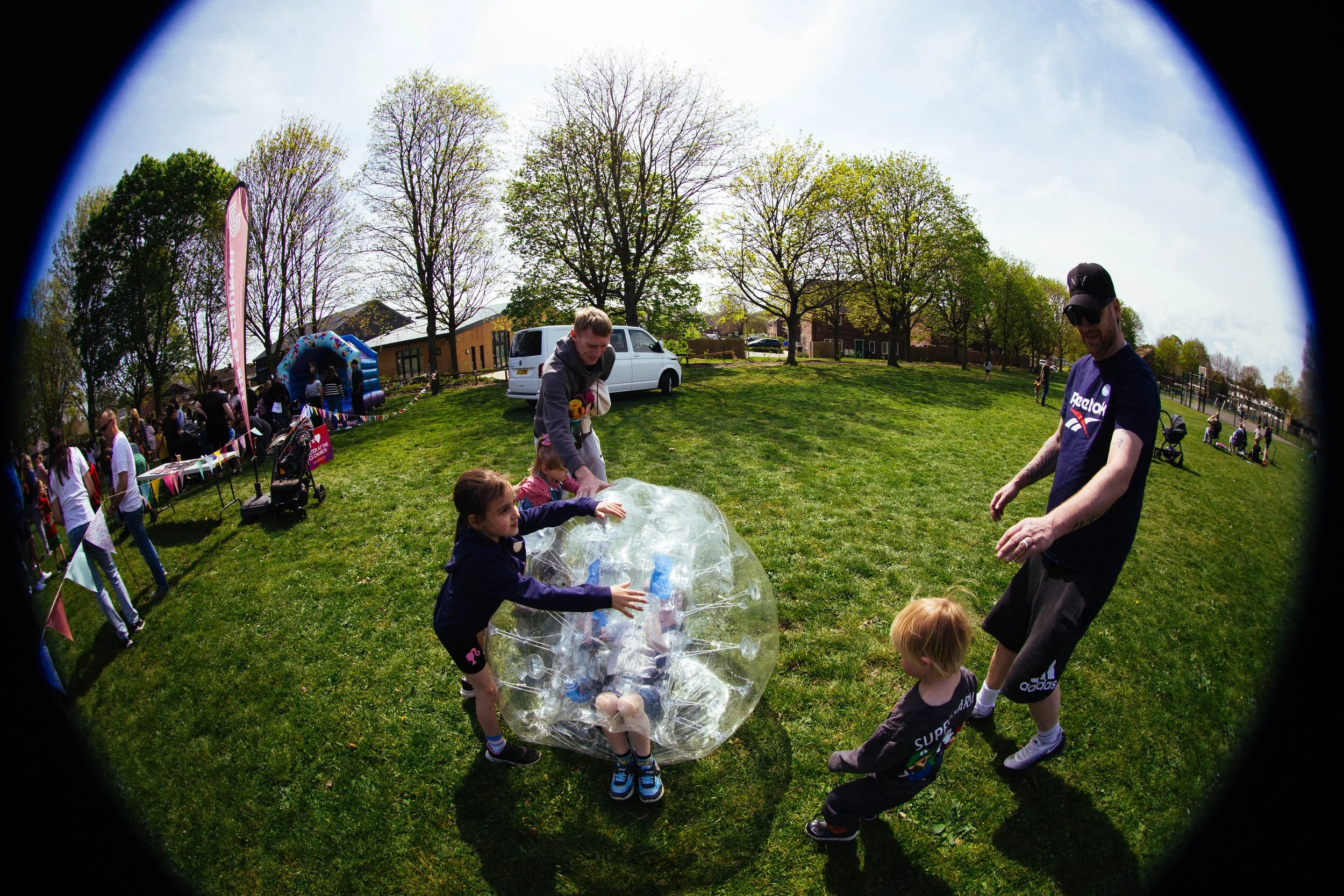 Children and adults at an outdoor event, with some children inside a large transparent inflatable ball, on a grassy field with trees, tents, and people in the background.