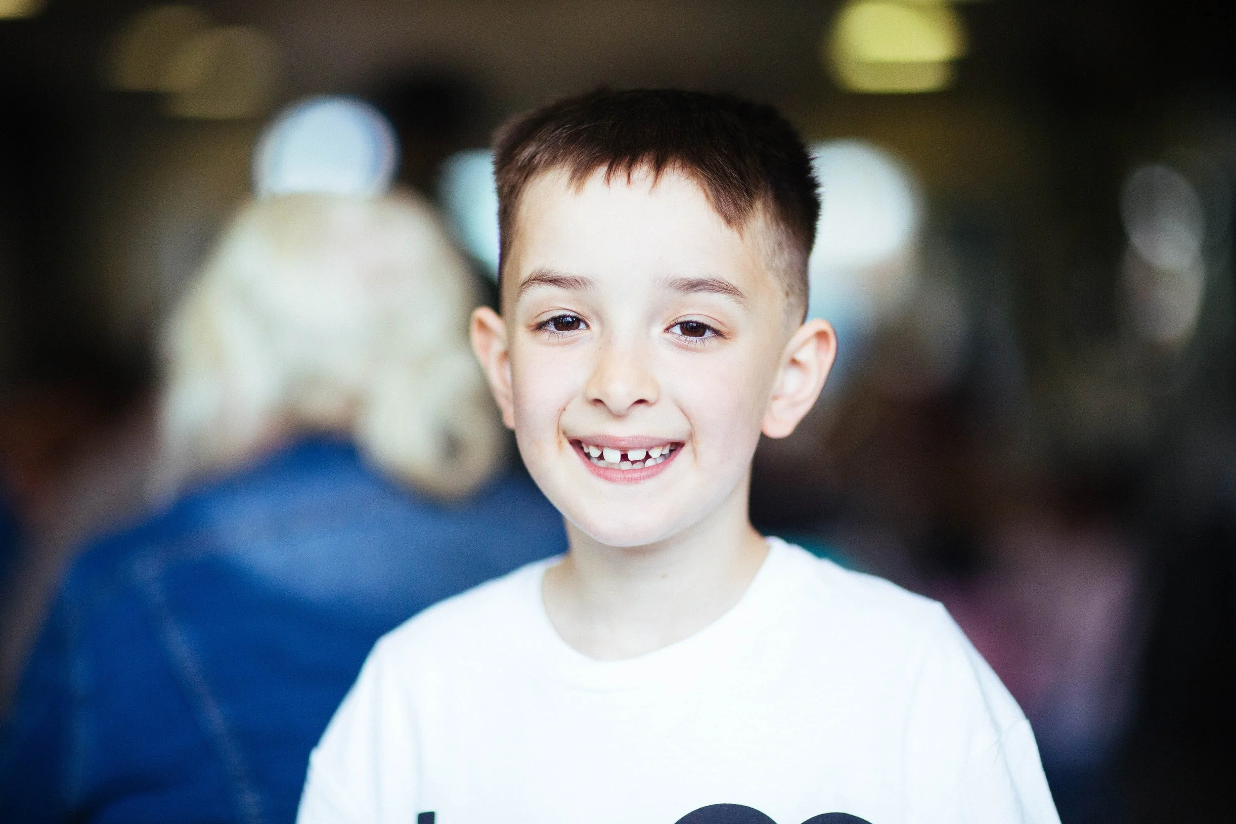 Smiling young boy with missing front teeth in a crowded indoor setting.
