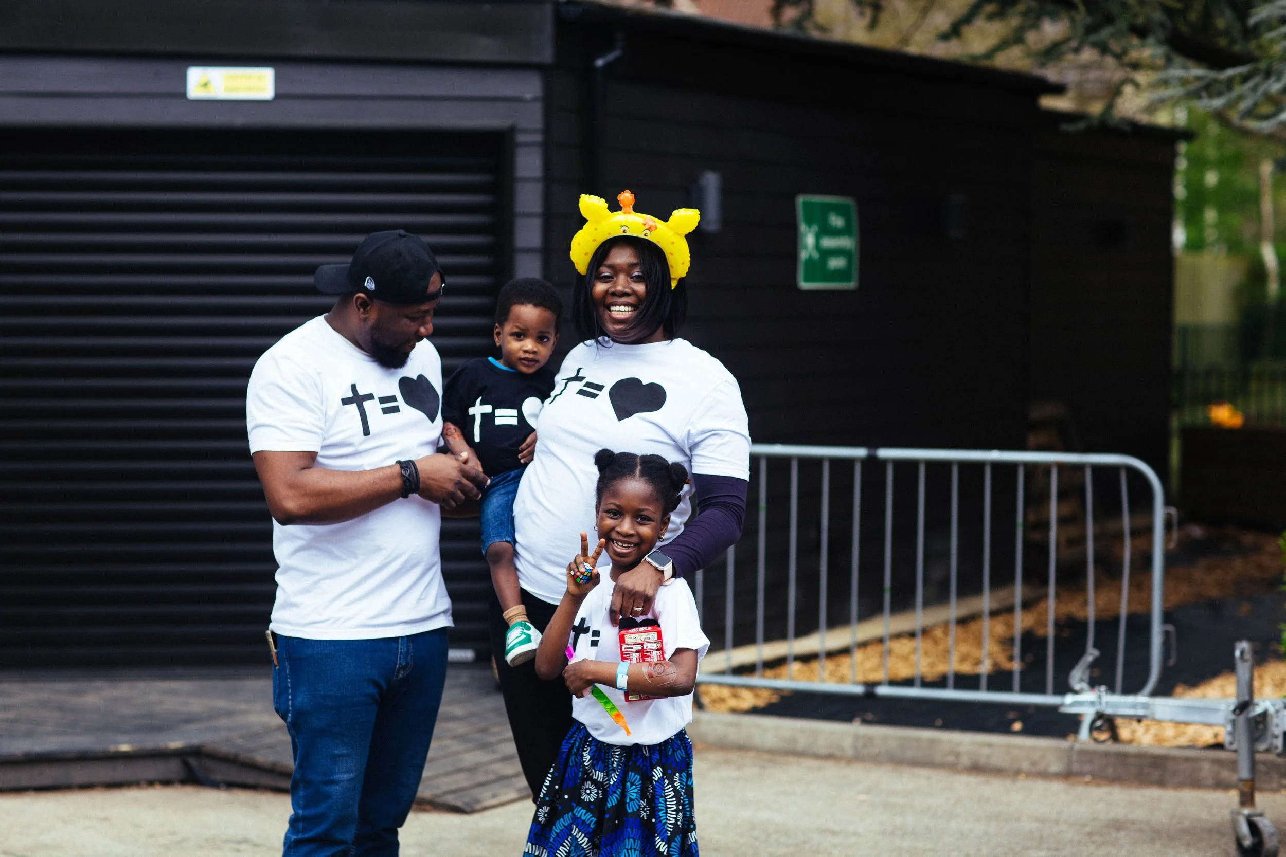 A family of four enjoying a day outdoors, all wearing matching white and black shirts with heart and math symbols, standing in front of a black building with a metal barrier nearby. The mother is smiling and wearing a yellow giraffe headband, while t