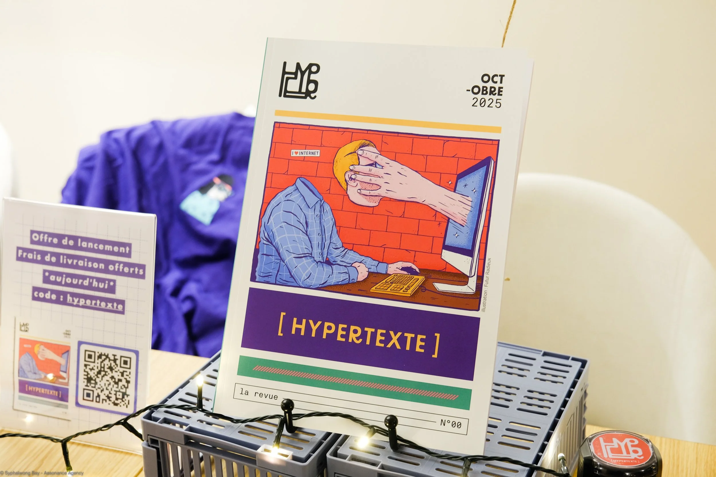 Une affiche promotionnelle pour une revue nommée [HYPERTEXTE], avec une illustration d'une personne assise devant un ordinateur dont une main énorme sort d'un écran pour couvrir son visage. La scène est dessinée dans un style coloré. La revue est dat