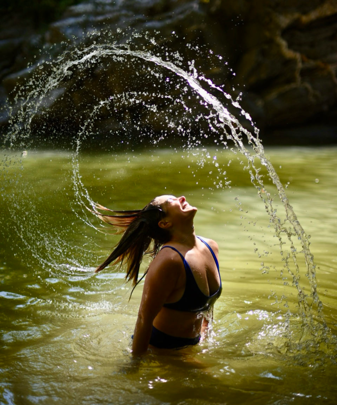 A photo of Charlene in a blue swimsuit throwing her head back in water, creating a splash in an outdoor setting with a rock overhang in the background.