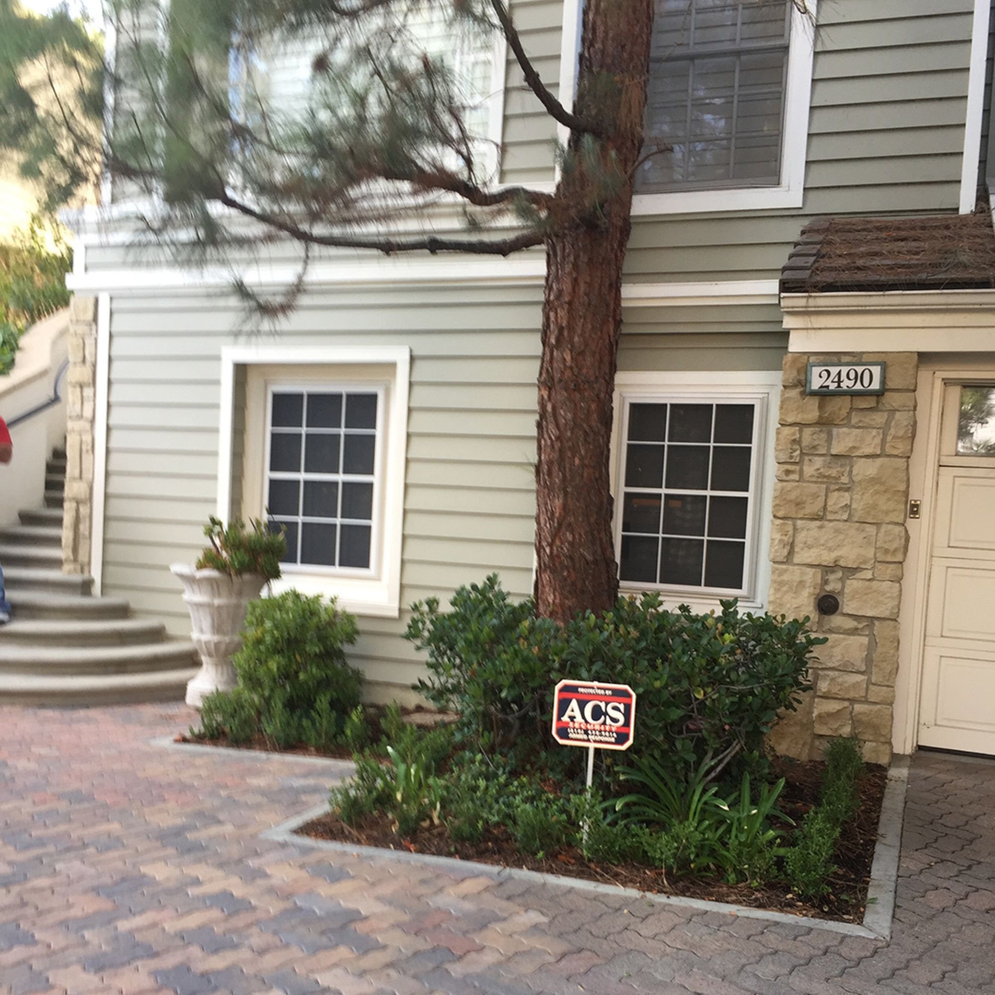Suburban house with gray siding, trees, shrubs, and a security sign in front.