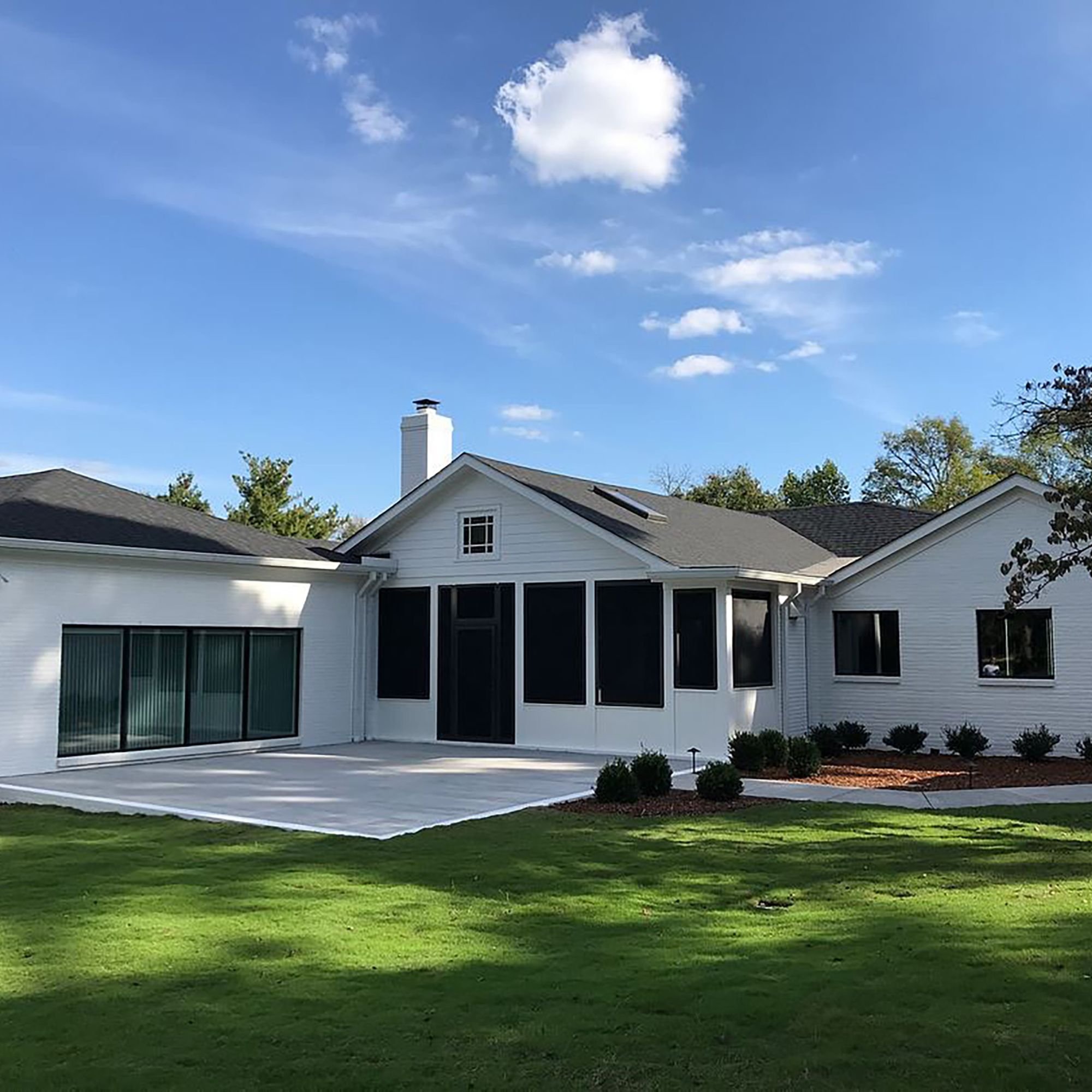 Modern white residential house with black trim, large windows, and a chimney, set against a blue sky with scattered clouds. The yard has neatly trimmed grass and shrubbery.