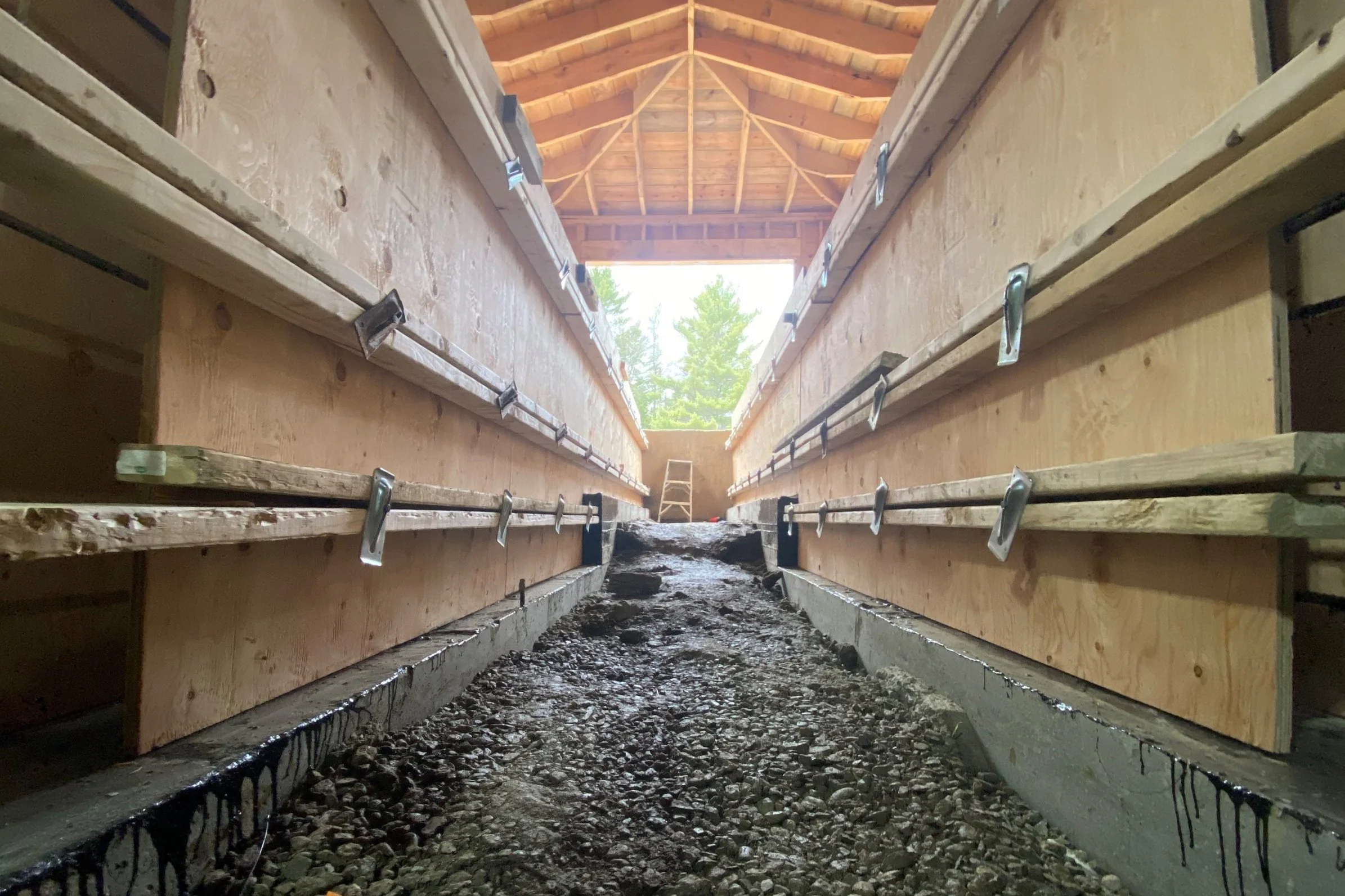 Construction site showing wooden walls and ceiling with metal brackets, rough ground with stones, and an opening allowing natural light in.