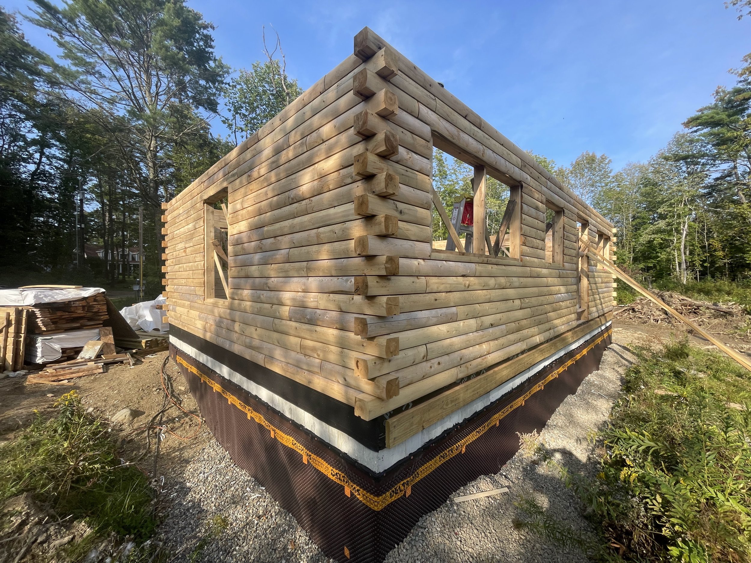 The image shows a wooden house under construction in a wooded area on a sunny day. The frame of the house is built with horizontal logs, with window openings visible, and construction material and tools are around the site.