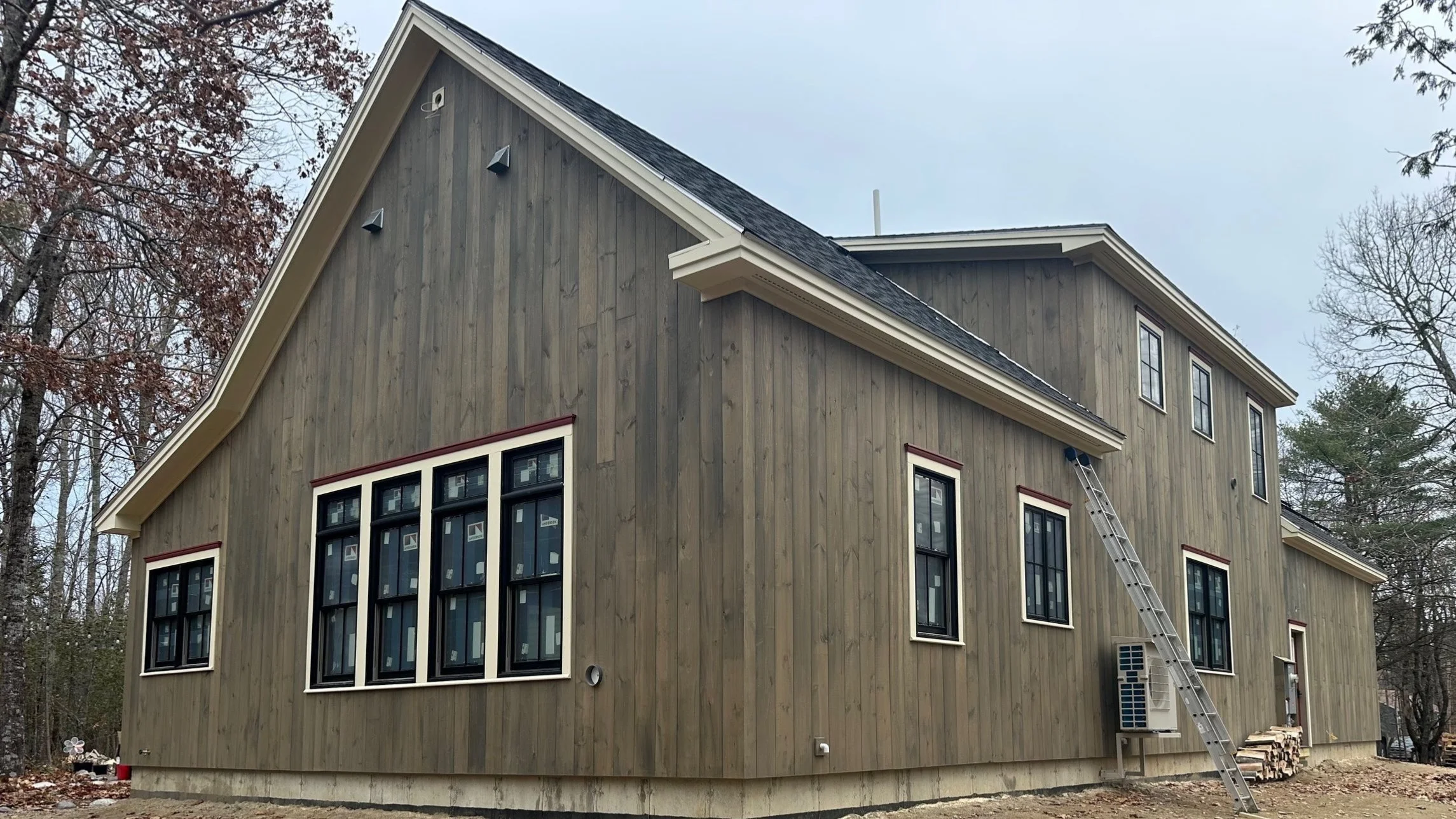 The image shows a new wooden house with multiple windows, a steeply pitched roof, and a surrounding wooded area. There is a ladder leaning against the house and a split firewood pile nearby.