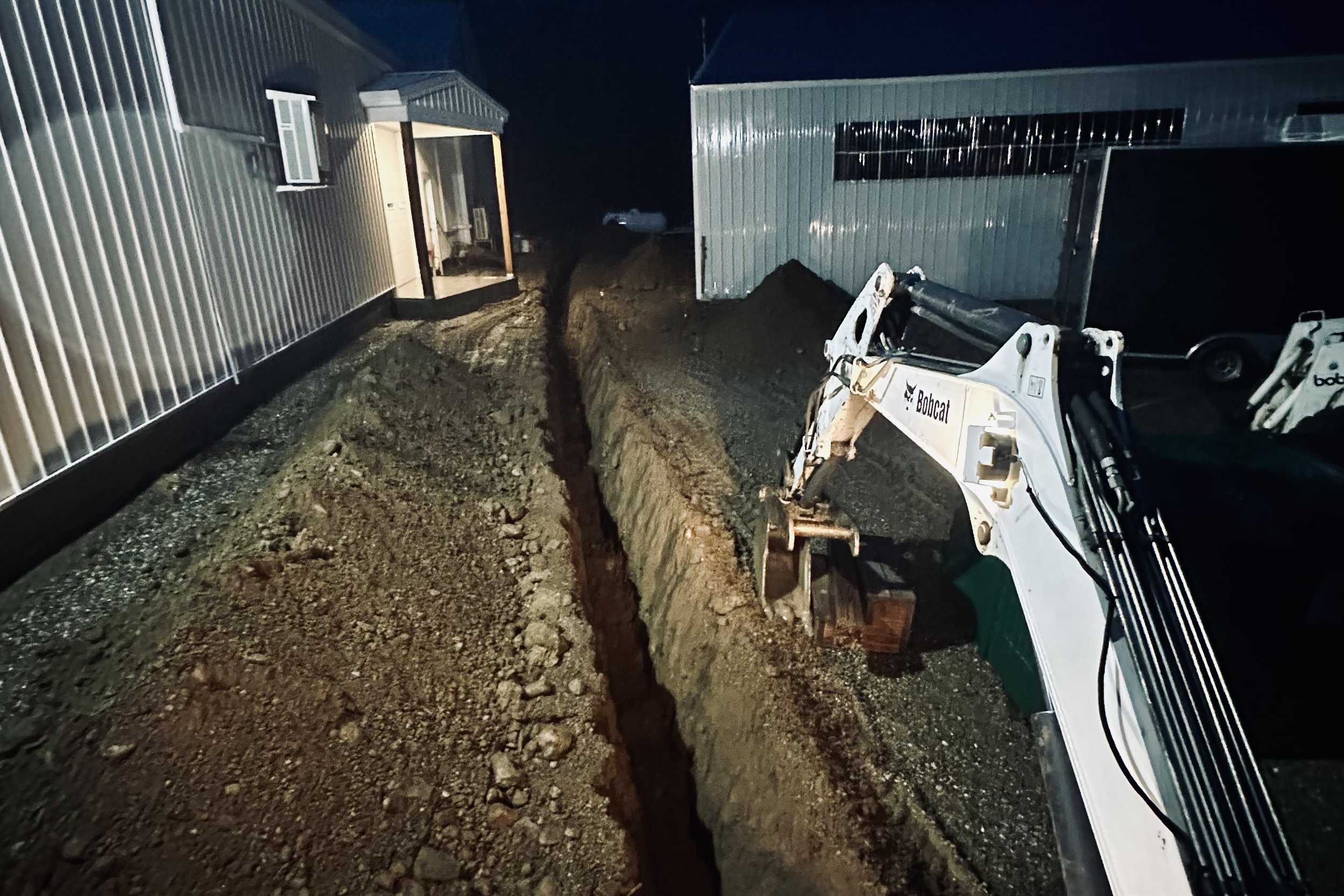 Nighttime construction site with excavator digging a trench between two metal buildings, dirt and rocks scattered around.