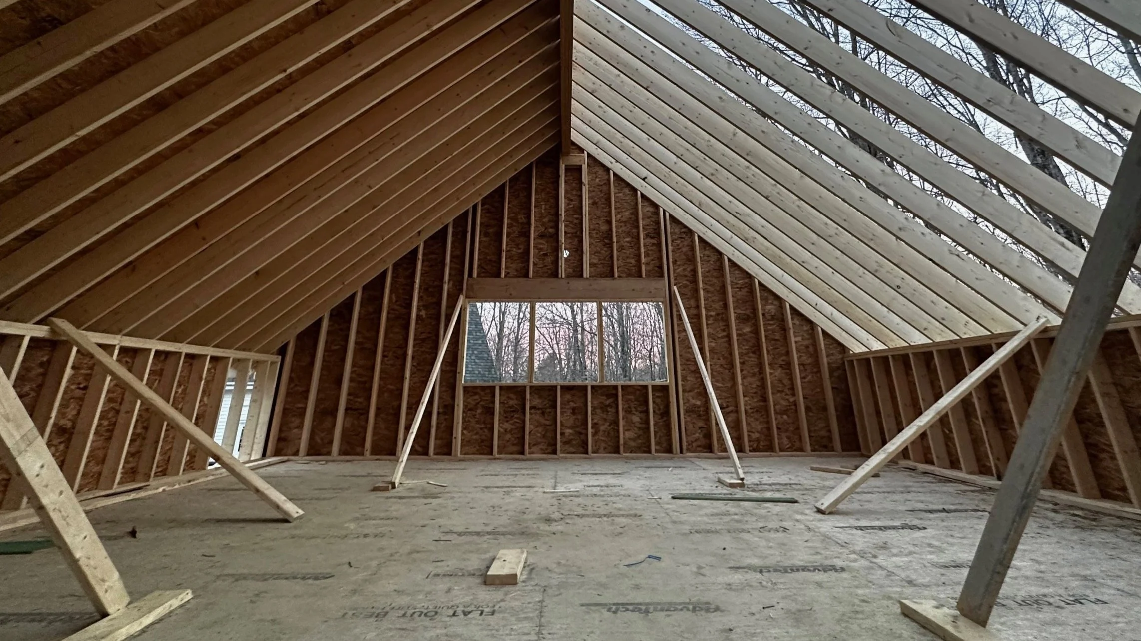 Interior view of a house under construction, showing unfinished wooden framing and roof structure with a window opening and a view of trees outside.