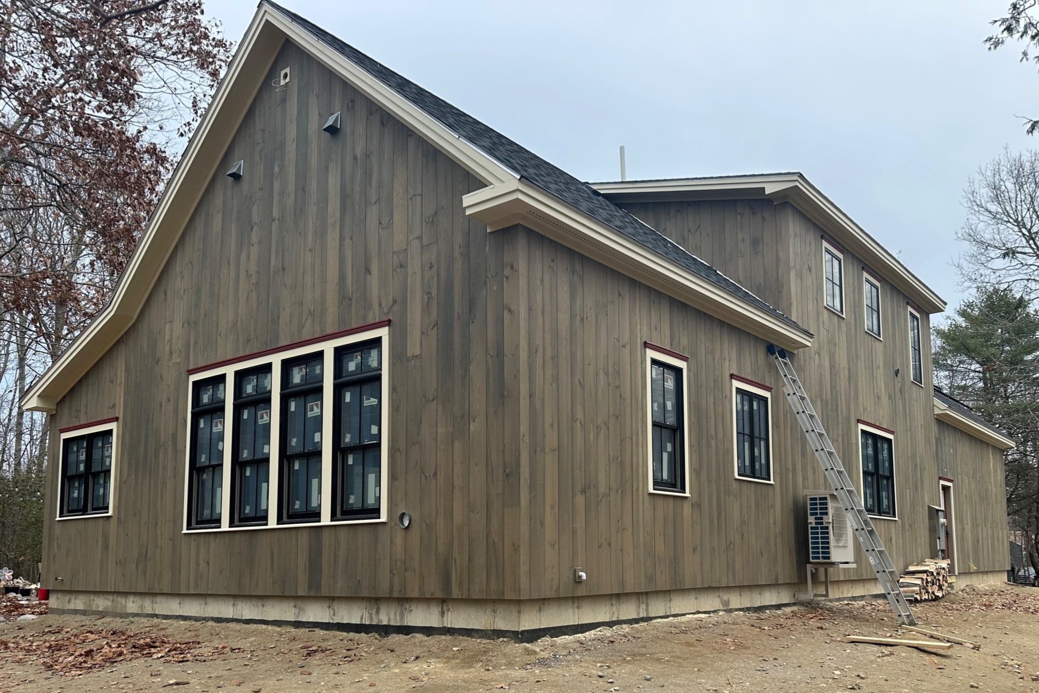 A two-story house under construction with gray wooden exterior siding, multiple large windows with black frames, a steep gable roof, and a ladder leaning against the side. There are trees in the background and construction materials on the ground.