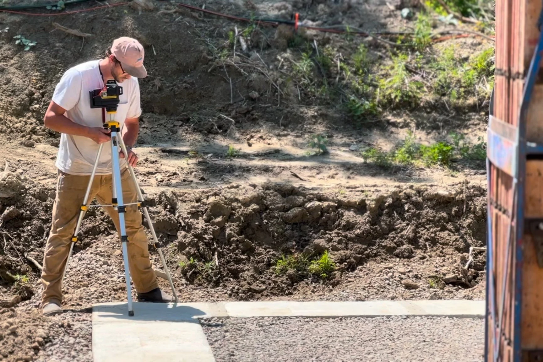 A man in a beige cap and tan pants using a surveyor tripod in an outdoor construction area.