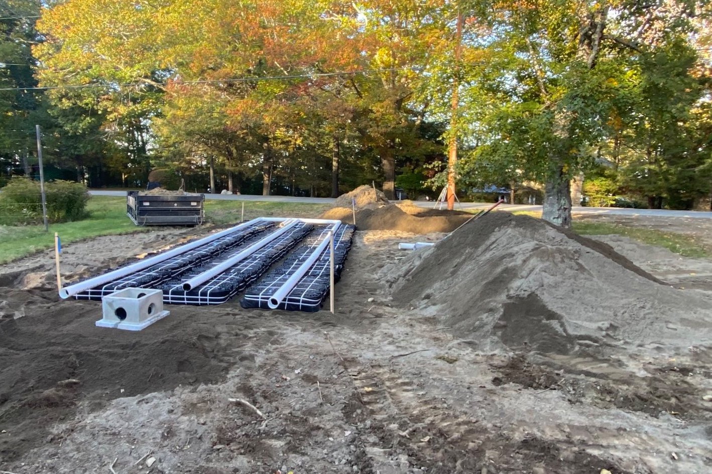 Construction site with piping and gravel piles, surrounded by trees with autumn leaves.
