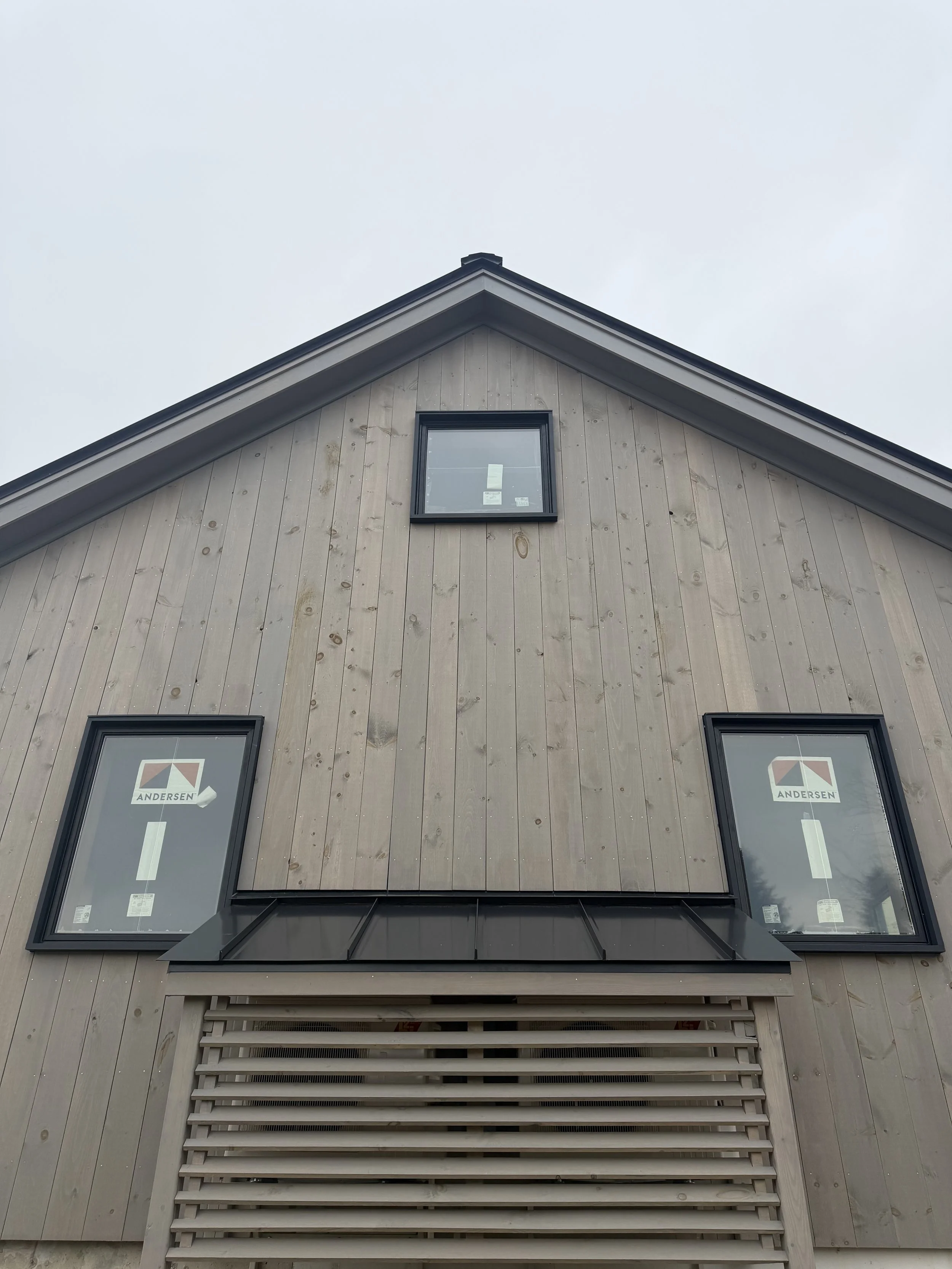 Close-up of a modern house's upper facade with beige wooden siding, three windows with black frames, and horizontal ventilation slats below the bottom window.