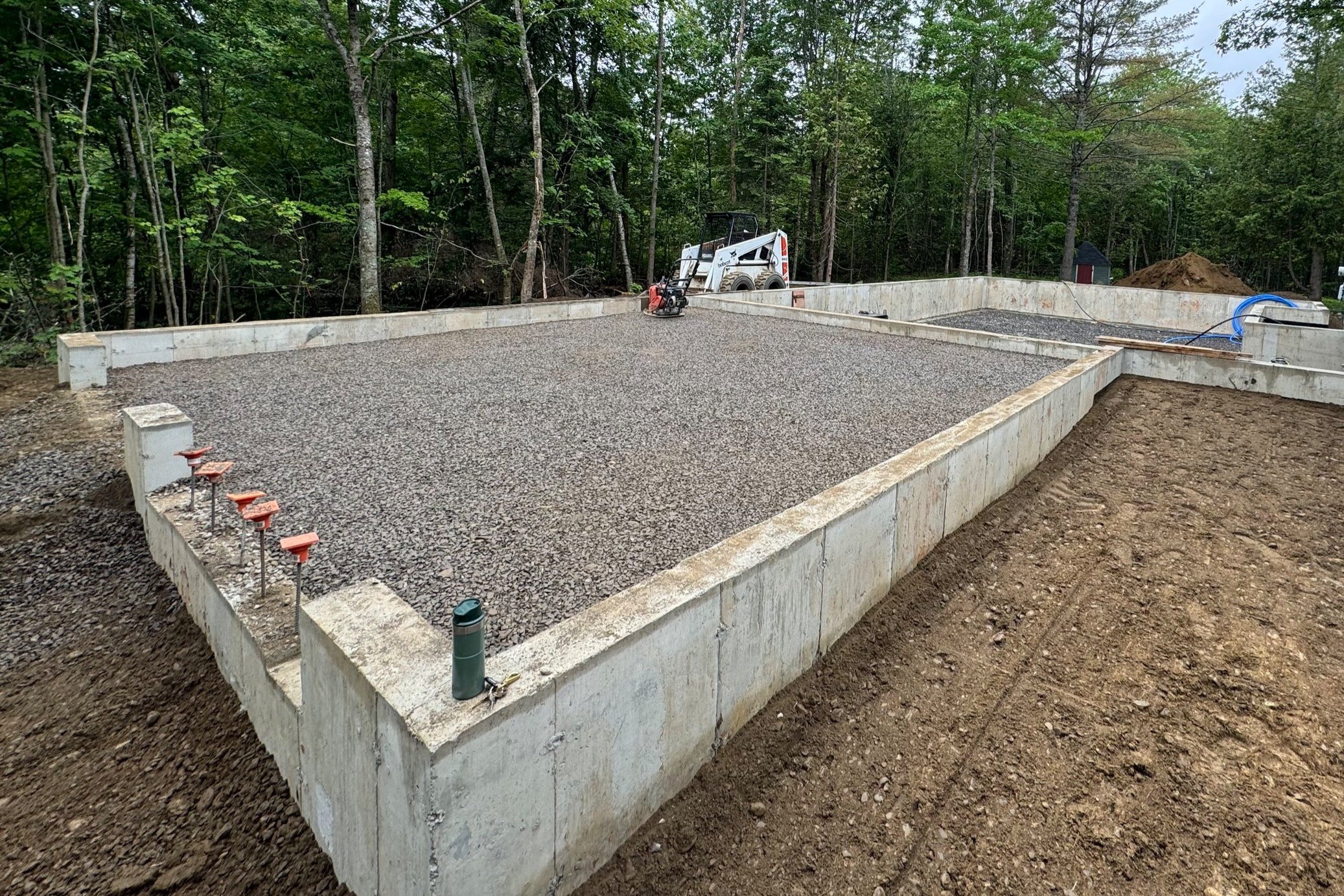 A construction site with a concrete foundation and gravel top, surrounded by forest trees and construction equipment, including a small skid-steer loader and a blue hose.