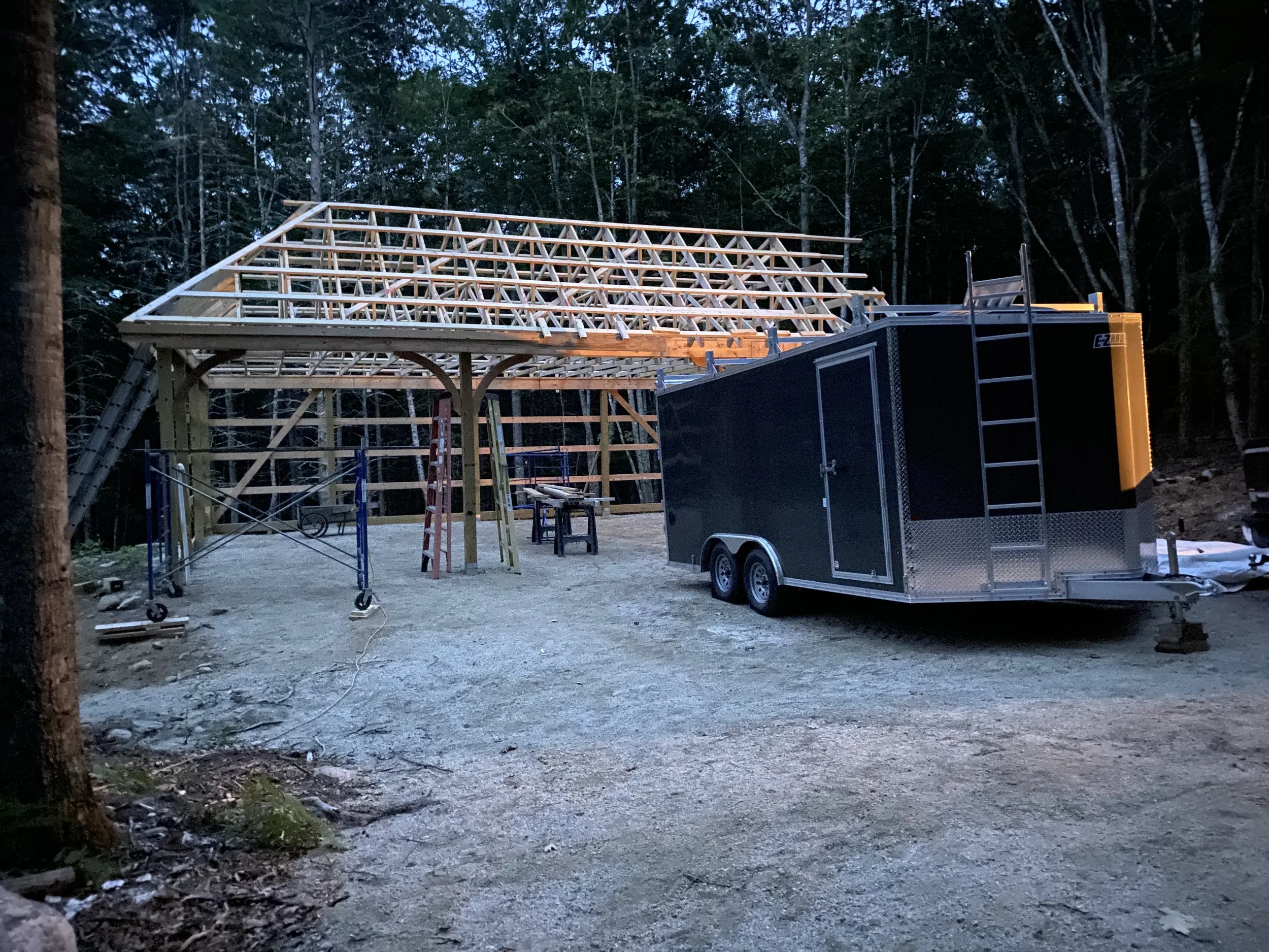A wooden roof frame is under construction on a small platform in a forest clearing during twilight, with construction tools and a trailer nearby.