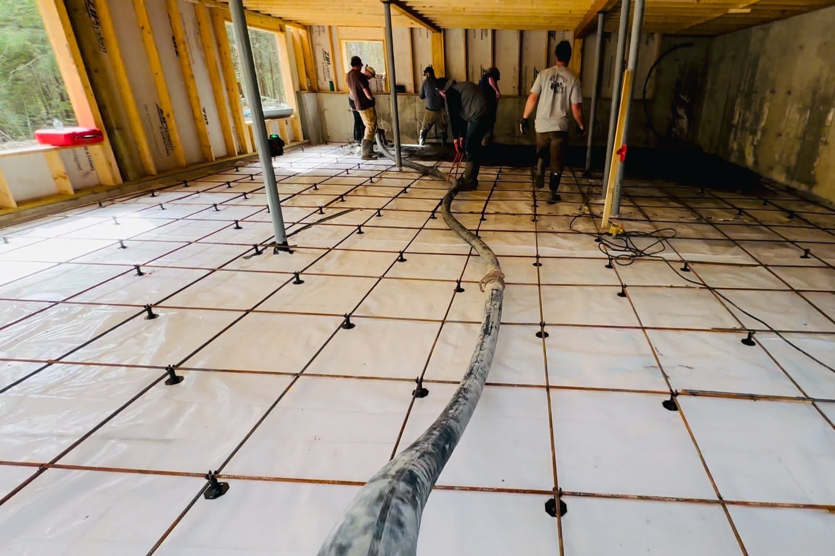 Workers pouring concrete over a heated wire mesh floor in a room under construction.