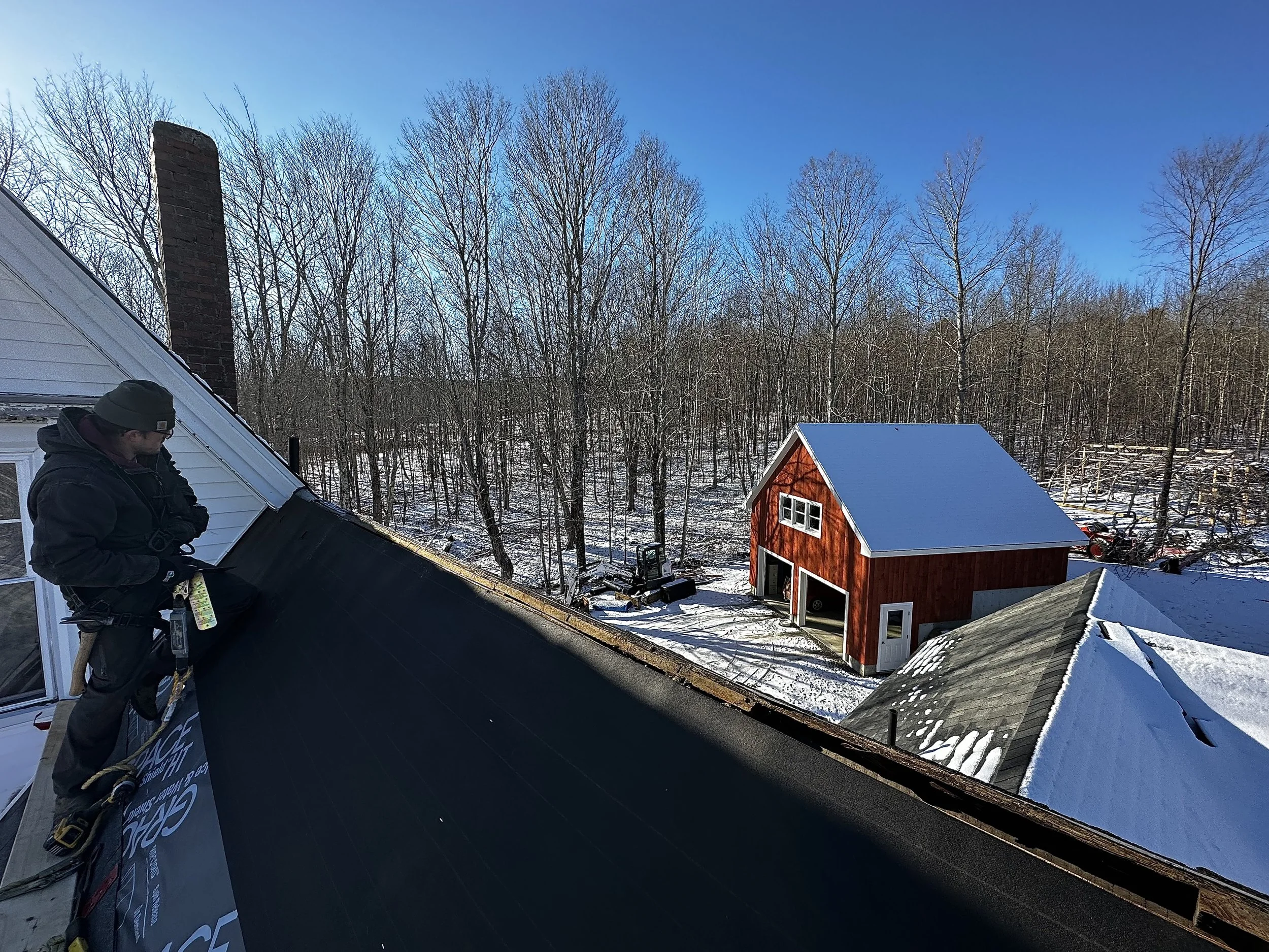 A man wearing a green beanie and black jacket working on a roof, installing new black shingles on a residential house with a snow-covered yard and a clear blue sky.