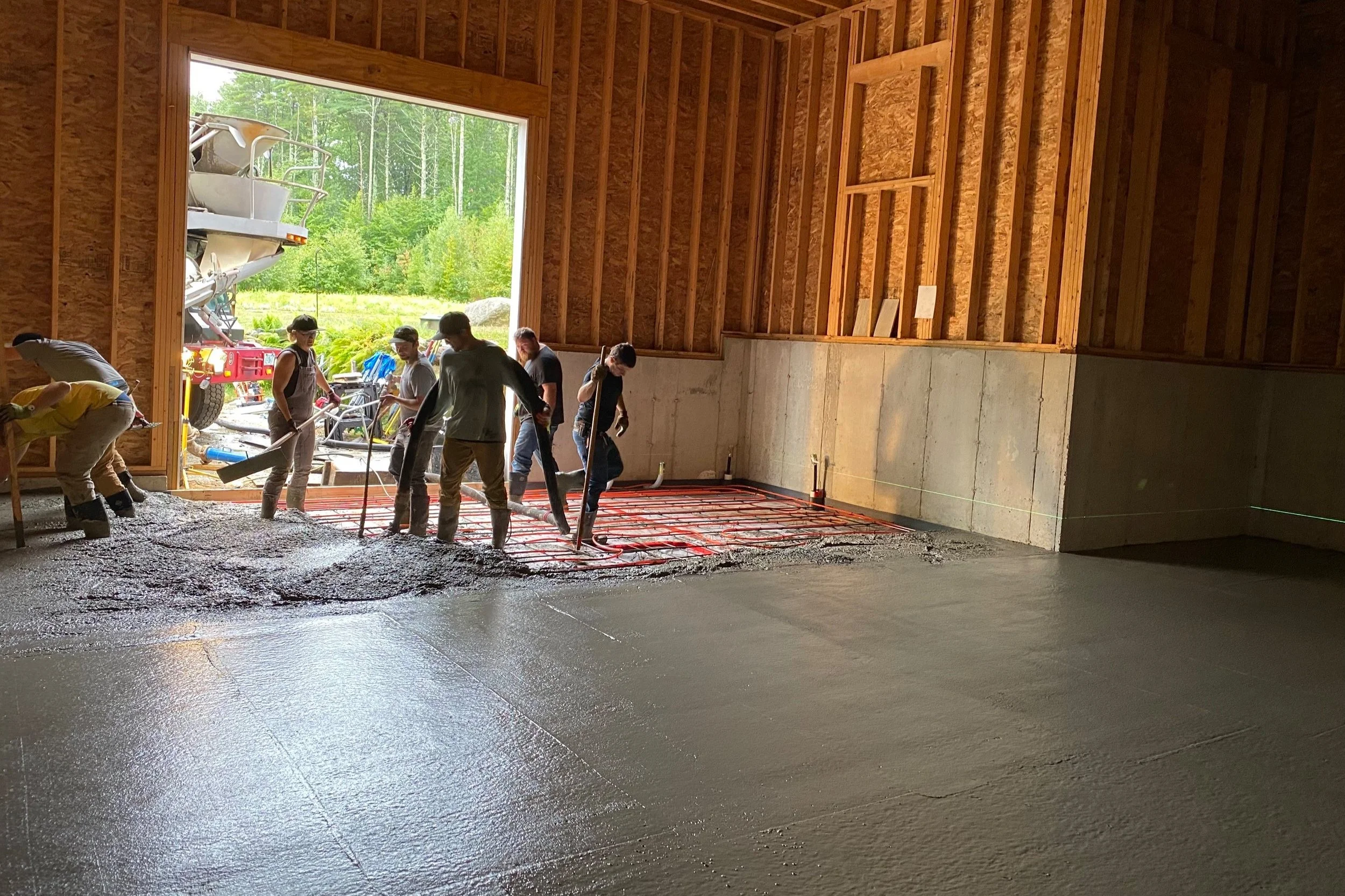 Workers pouring and leveling concrete in the basement of a house under construction, with a large open garage door showing a forested area outside.