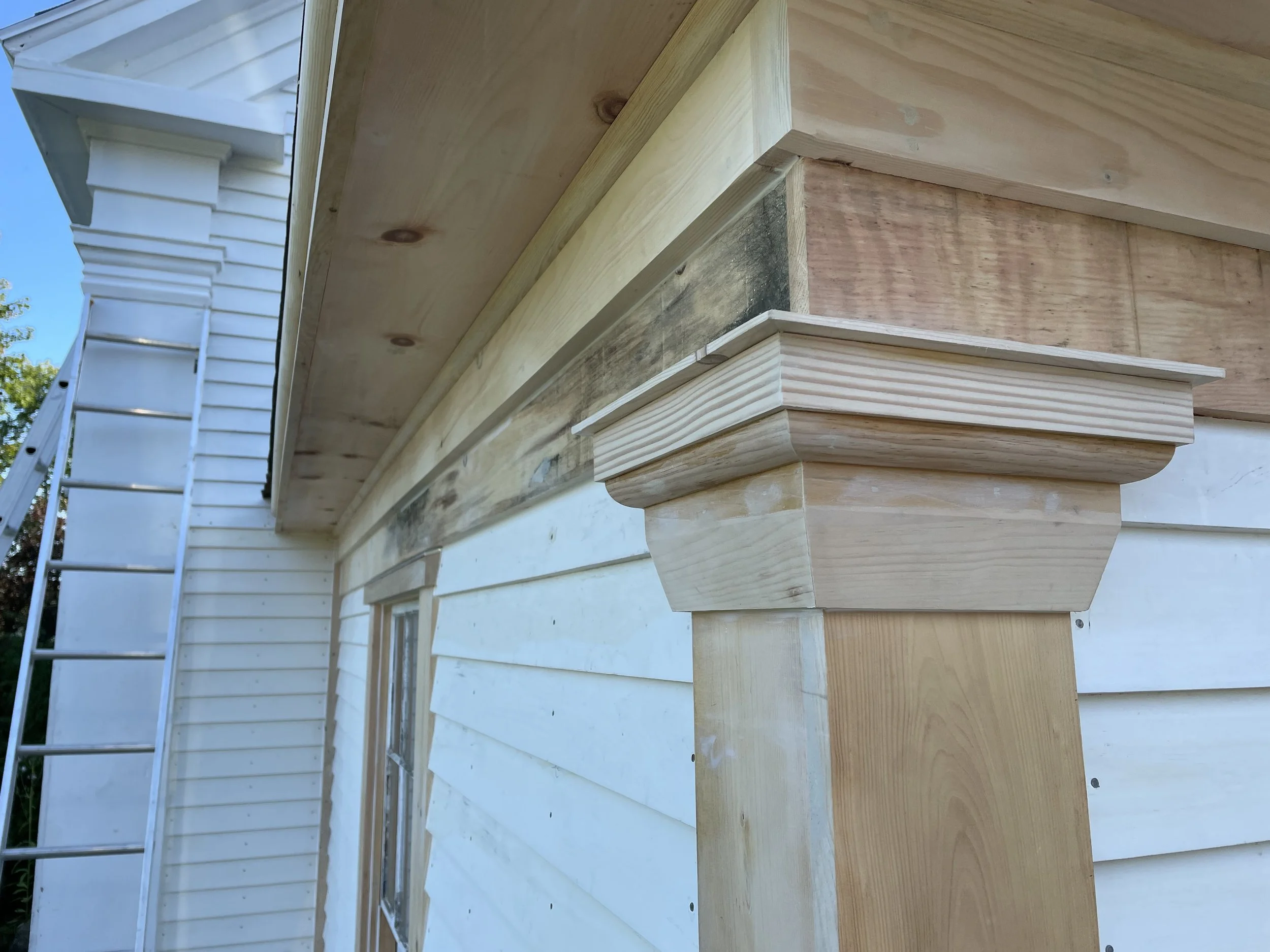 Close-up view of wooden architectural trim and molding on a house exterior with a section of white siding and a ladder in the background under a blue sky.