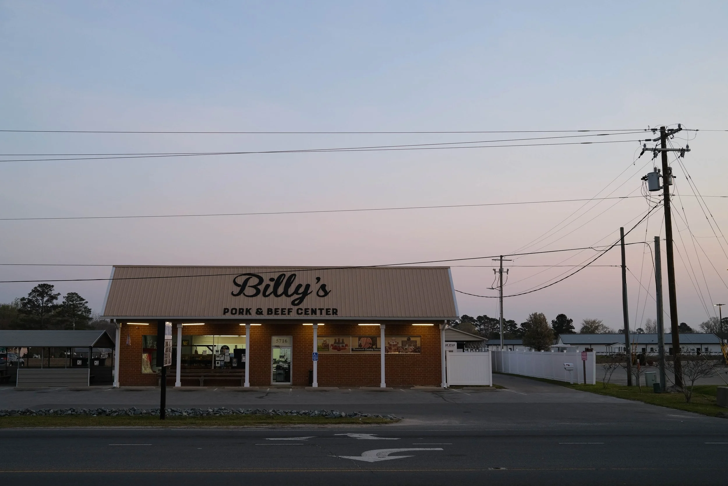 Brick and aluminum siding storefront on highway. The building sign says "Billy's Pork and Beef Products." A purple and pink dawn sky in the background. Wallace, North Carolina.