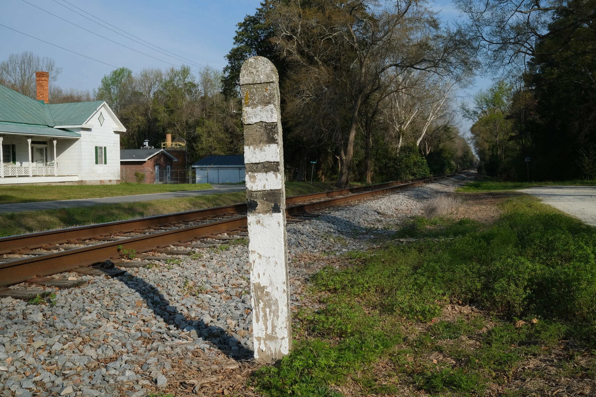 White and black striped railroad sign beside track in Magnolia, North Carolina. In the background stands a white house with green metal roof from early 1900s.