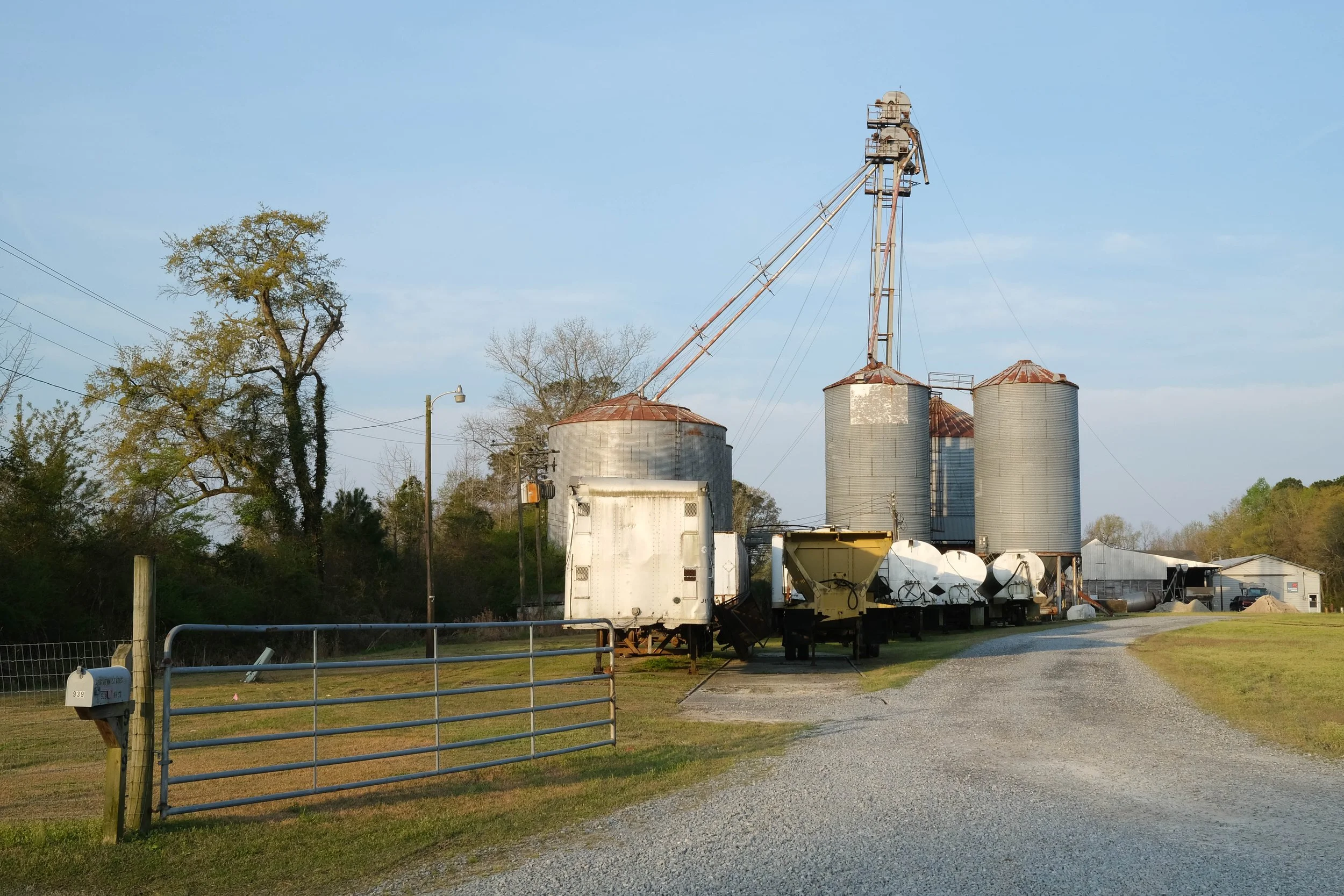 Four aluminum silo feed mill with many outbuildings and truck trailers in railyard beside railroad track. Wallace, North Carolina.