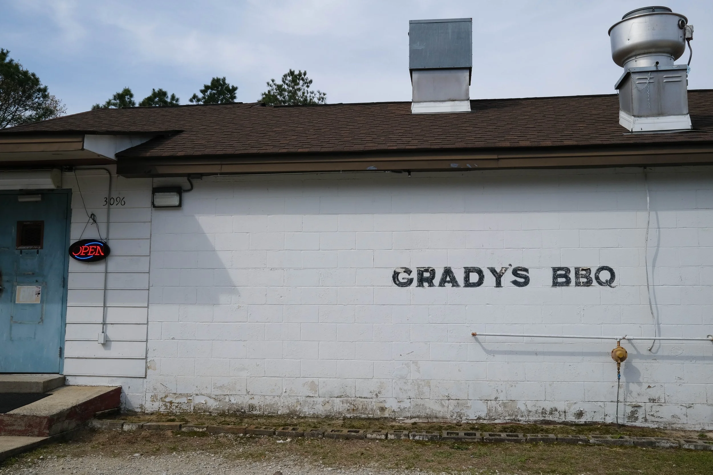 Cinderblock building painted white with blue door. Blinking lit sign that says "Open." Painted on the side of the building is "Grady's BBQ." Dudley, North Carolina.