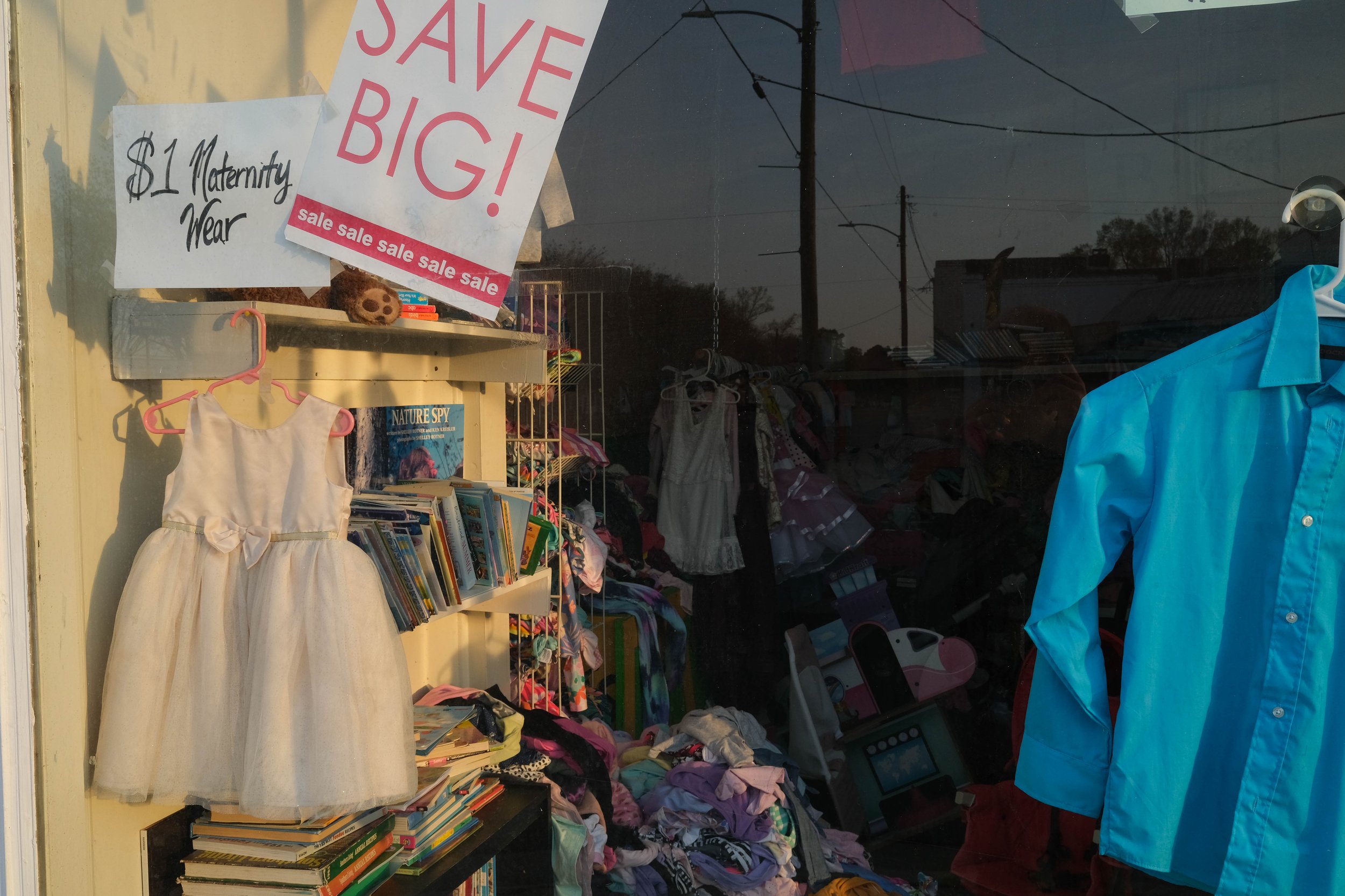 Storefront window in downtown Wallace, North Carolina. In window is sign that says "Save Big!" Another says "$1 Maternity Wear." A child's blue shirt and a little girls' Easter dress hangs in the window.