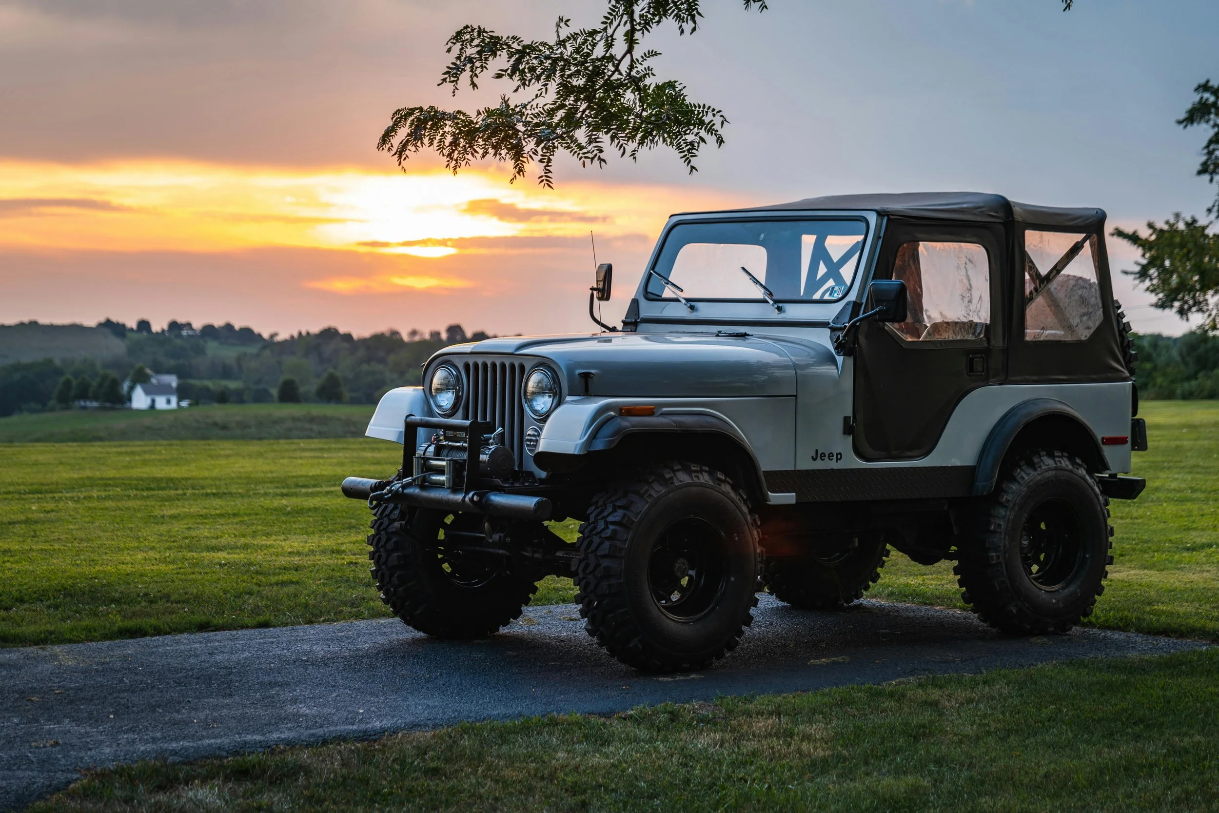 A gray Jeep with large off-road tires parked on a grassy area at sunset, with a scenic rural landscape in the background.