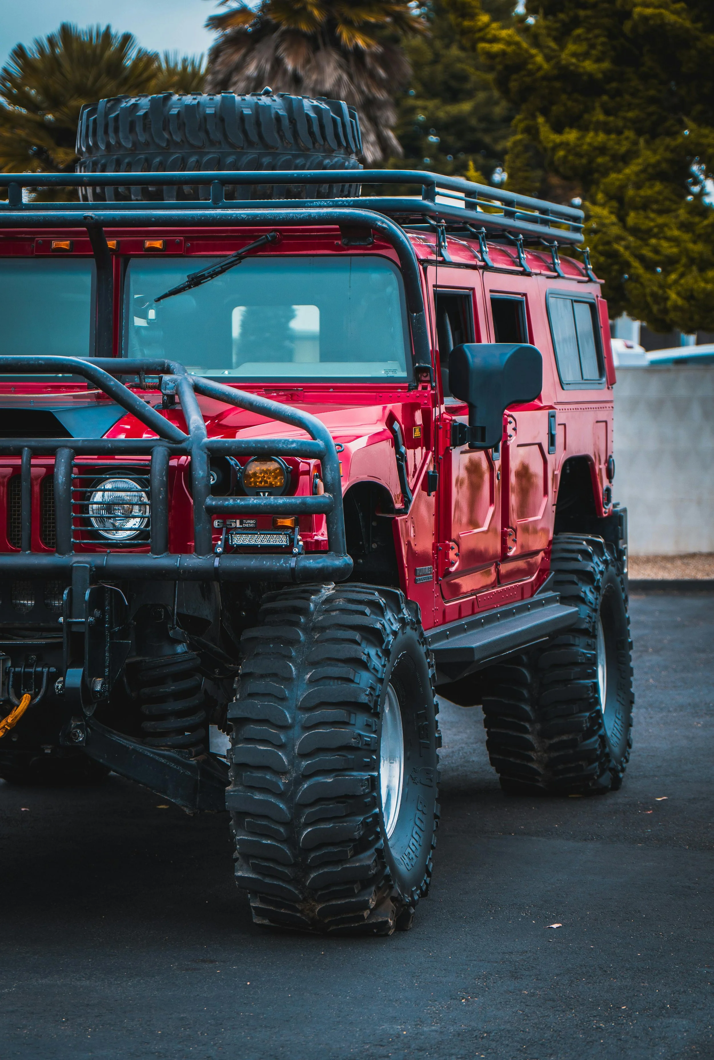 Red off-road vehicle with large tires, roof rack, and front bumper guard parked on asphalt, with trees in the background.