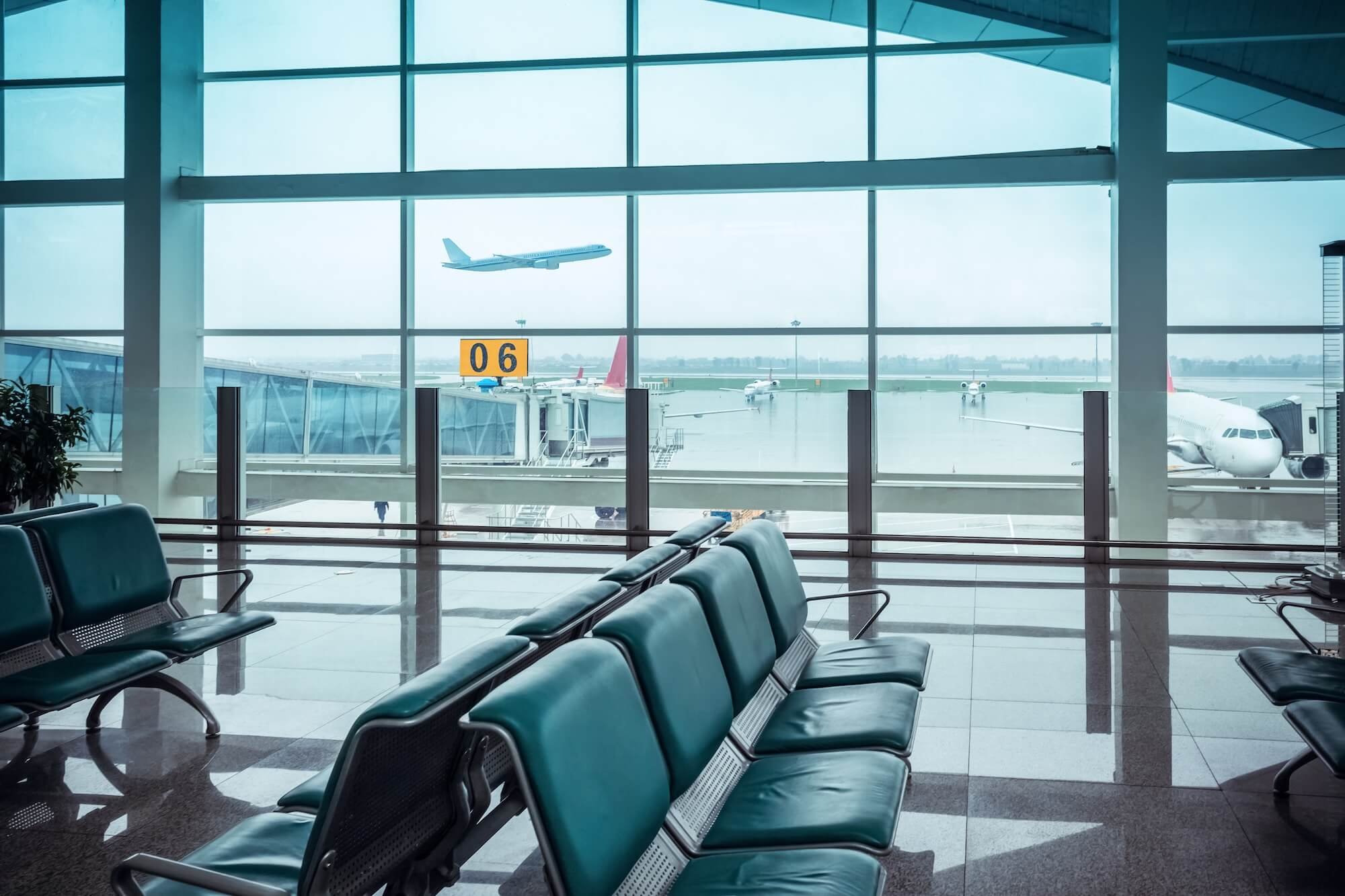 View of an airport terminal waiting area with teal chairs, large glass windows, a yellow gate number sign reading '06', and airplanes visible outside, including one taking off.