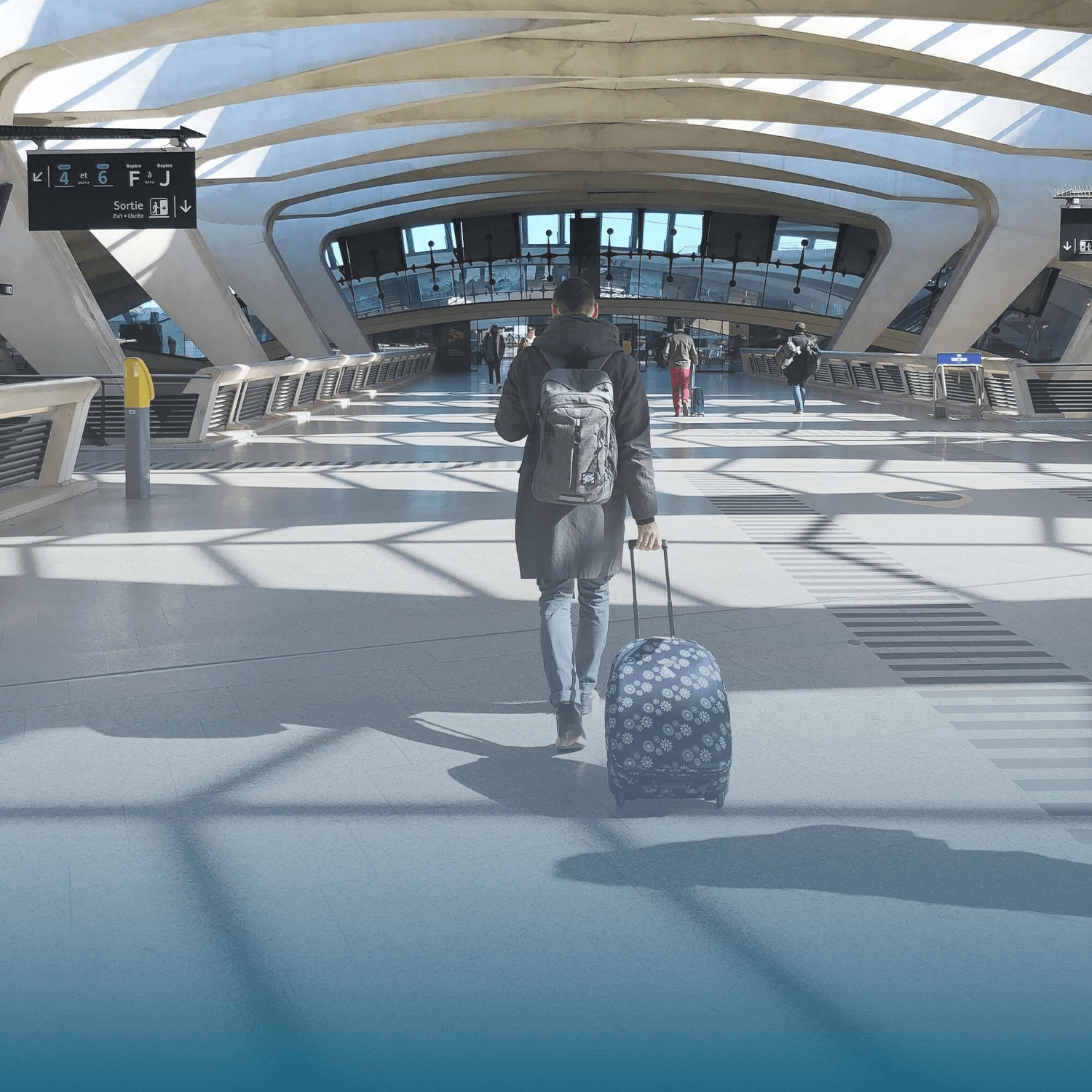 A man walking with a rolling suitcase in an airport terminal, with other travelers in the background and modern architectural design with curved beams and large glass windows.