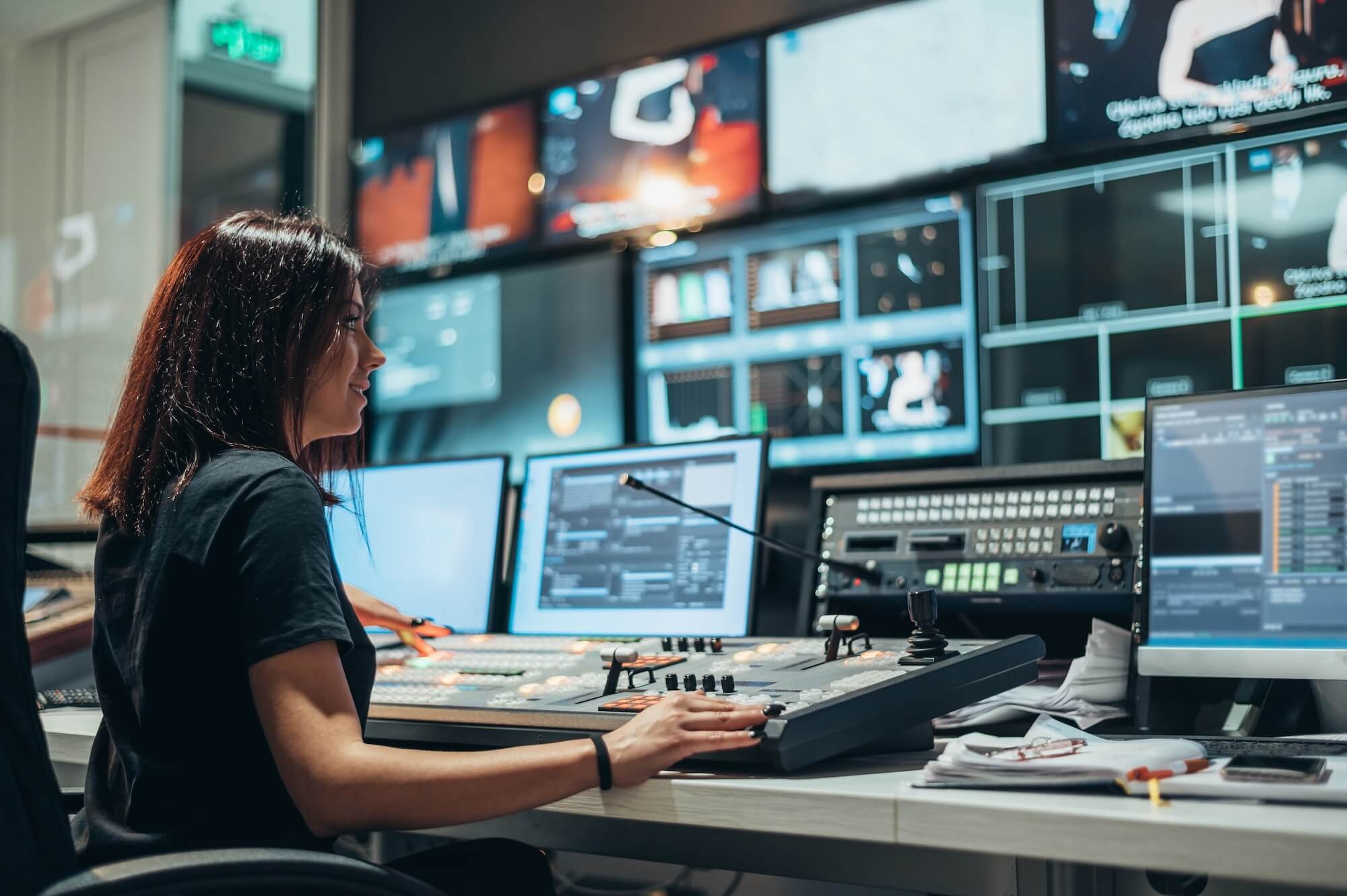 A woman operating a video editing console in a control room with multiple monitors displaying video feeds.