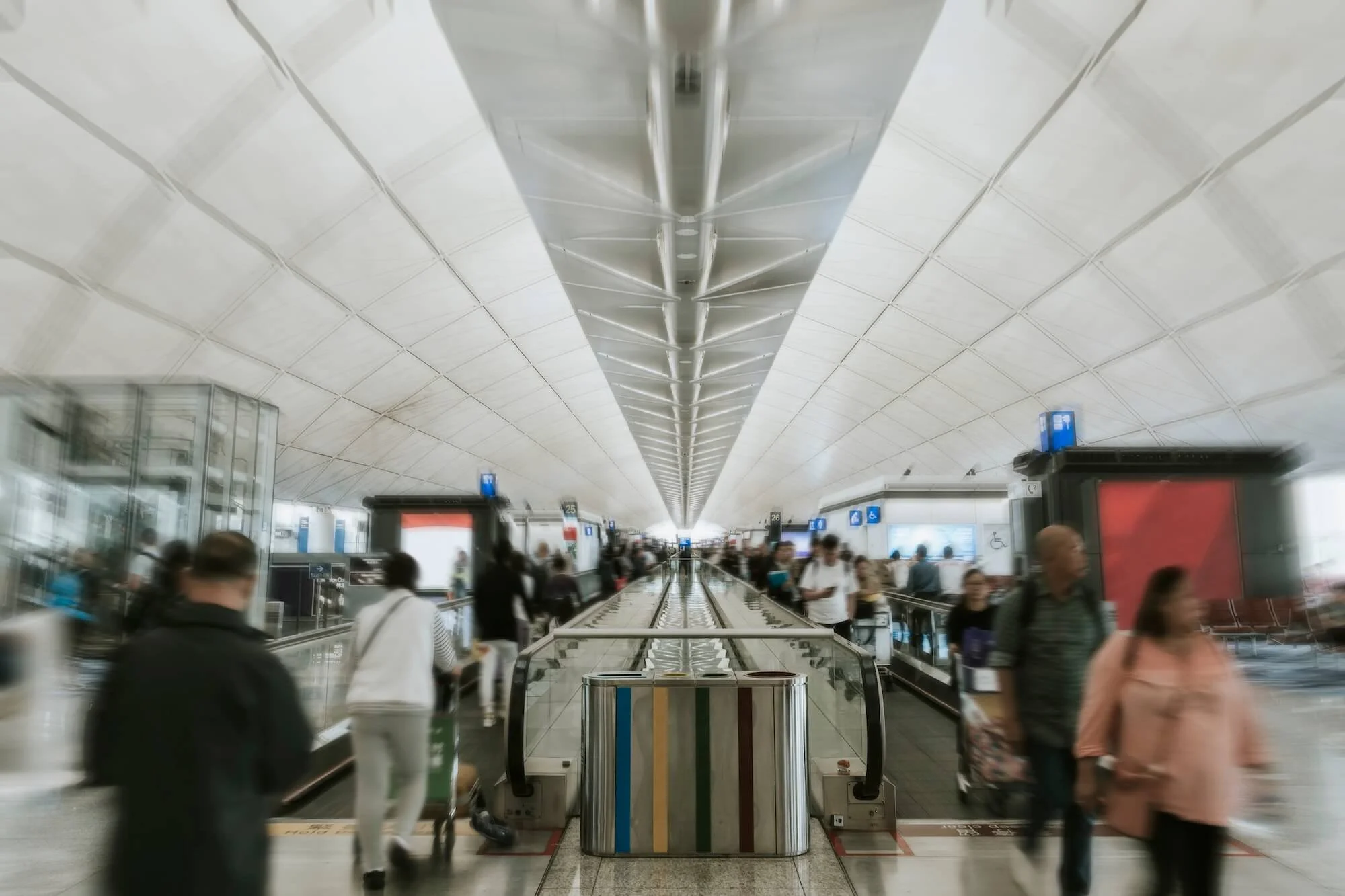 Interior of an airport terminal with a high arched ceiling, busy with travelers walking and standing near boarding gates and check-in counters.